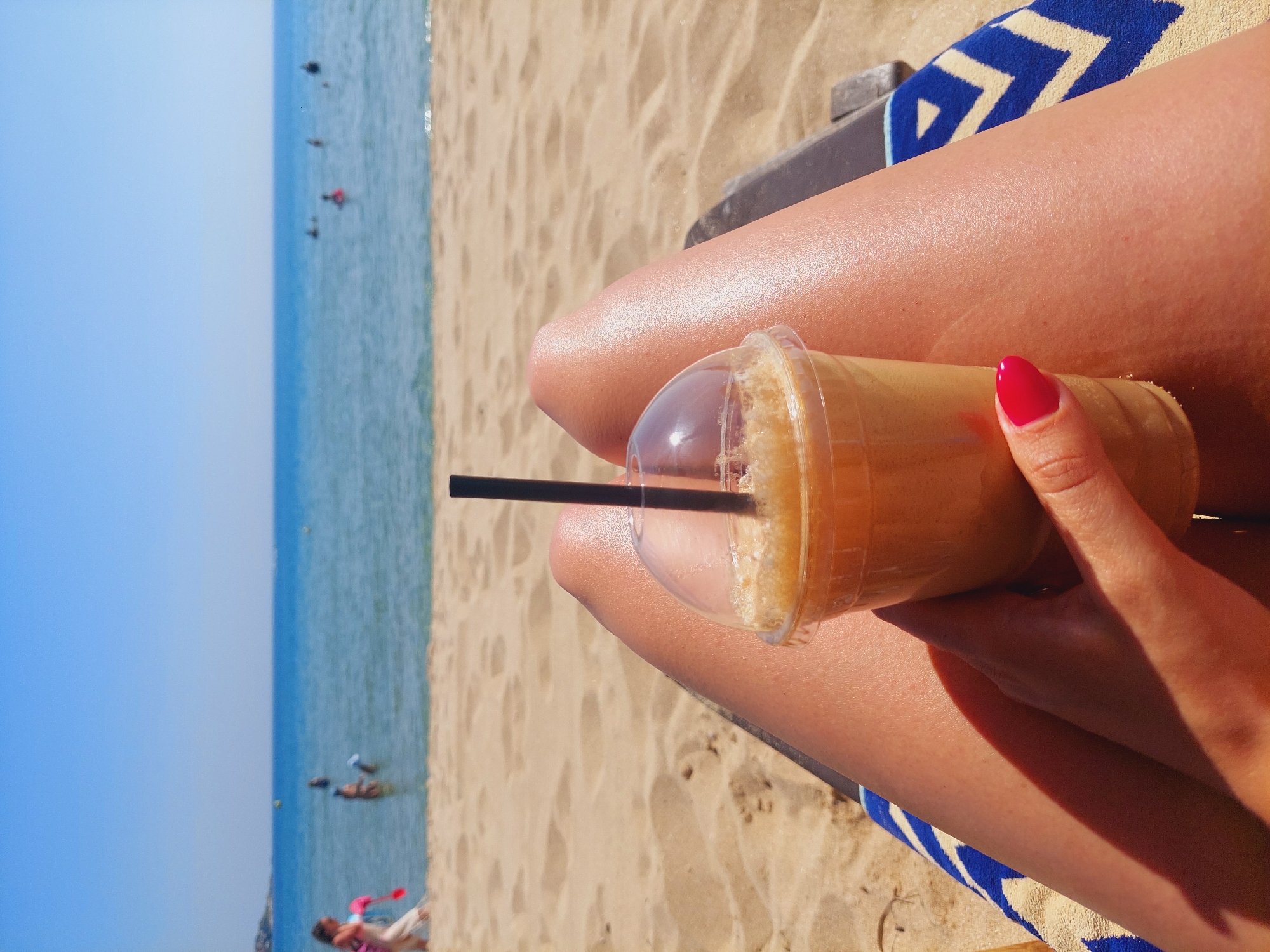 The image shows a first-person perspective of a person holding a takeaway coffee cup with a straw, with their legs propped up on a beach chair covered with a blue and white striped towel. The background is a sandy beach with the sea and a clear blue sky. There are a few people in the water, appearing to be swimming or wading. The coffee cup is in focus and is held in a right hand with bright red nail polish. The lighting suggests it is a sunny day. The location is identified as a beach in Sozopol, Bulgaria, which is a coastal town on the Black Sea.