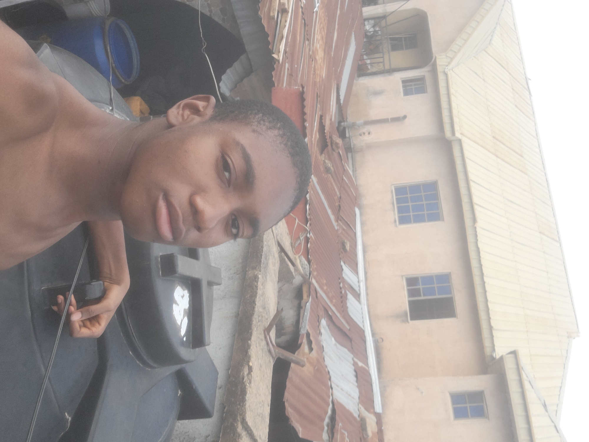 A young, shirtless man is taking a selfie. He is standing near a large, black plastic water tank. Behind him, there is a building with a weathered, corrugated metal roof and an adjacent, multi-story building with tan-colored exterior walls and windows. The location appears to be in Jos, Nigeria.