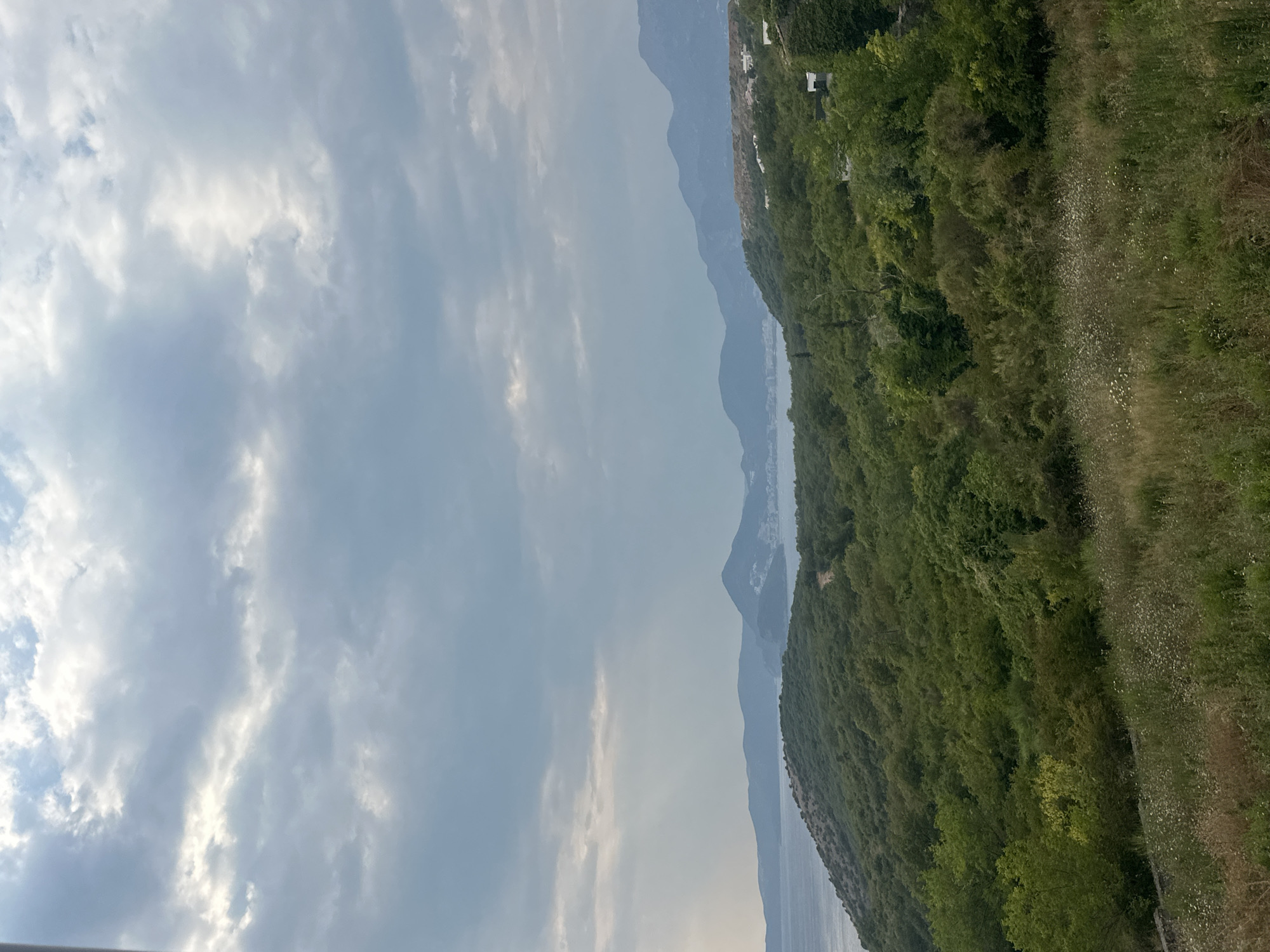 The image captures a panoramic view of the landscape around Drobnići, Montenegro. In the foreground, a grassy meadow with scattered wildflowers slopes down towards a dense forest covering the hillside. The forest is composed of various shades of green deciduous trees and shrubs. Beyond the forest, a body of water, likely the Adriatic Sea, stretches towards a series of hazy mountains in the distance. The sky is overcast with dramatic, textured clouds, allowing patches of sunlight to break through and cast rays onto the scene. A faint outline of buildings can be seen nestled along the coastline in the far distance, suggesting a coastal settlement. The overall impression is one of a natural, rugged landscape under a dynamic sky. There are no people or animals visible in the image.