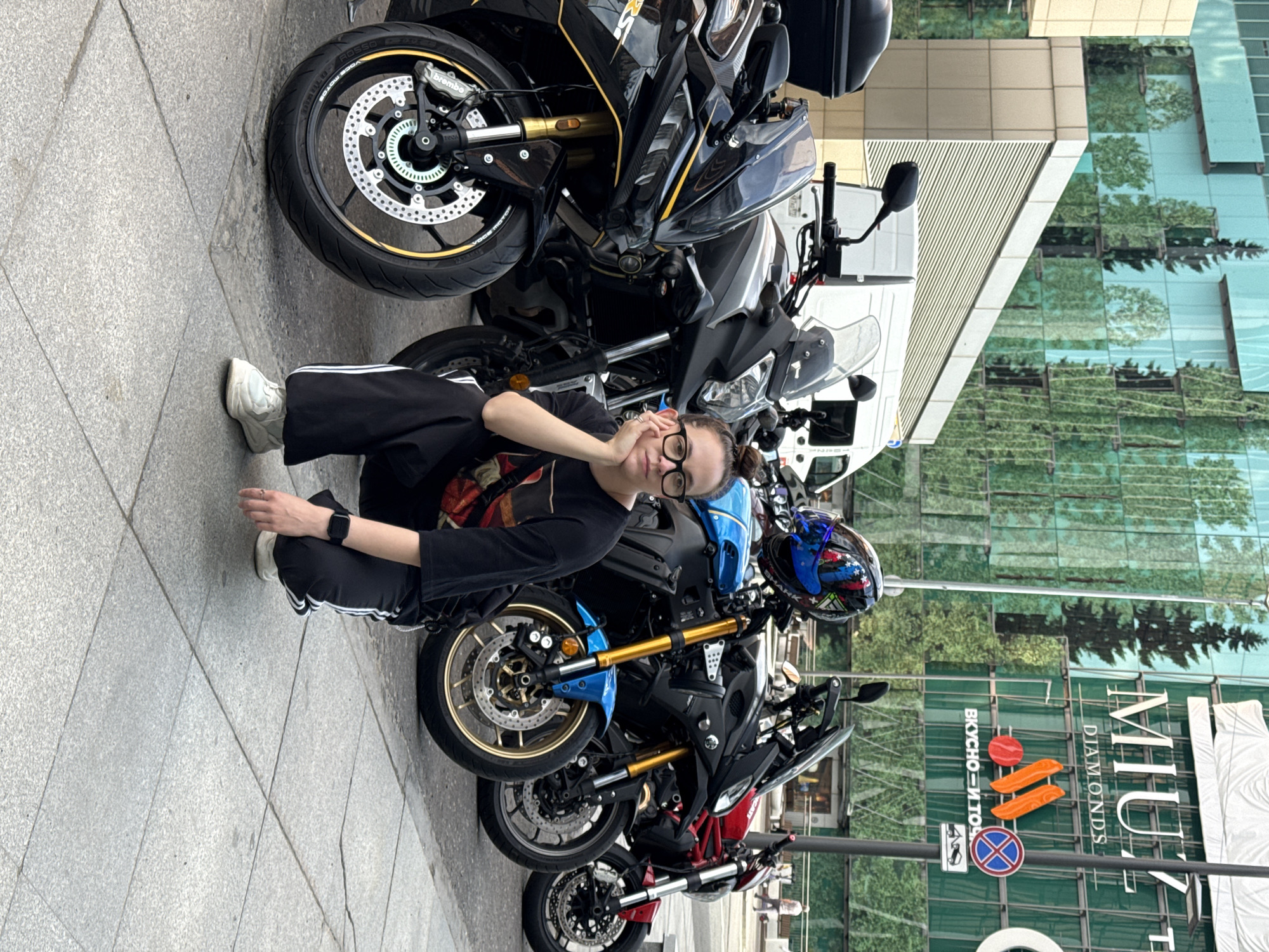 A young woman with glasses is crouching on a sidewalk in front of a row of motorcycles. She is wearing a black shirt and black pants with white stripes. The motorcycles are parked in front of a large building with a glass facade, reflecting trees and the sky. The building has the words "MIUZ DIAMONDS" and other Russian text visible. The location is identified as Khimki, Russia.