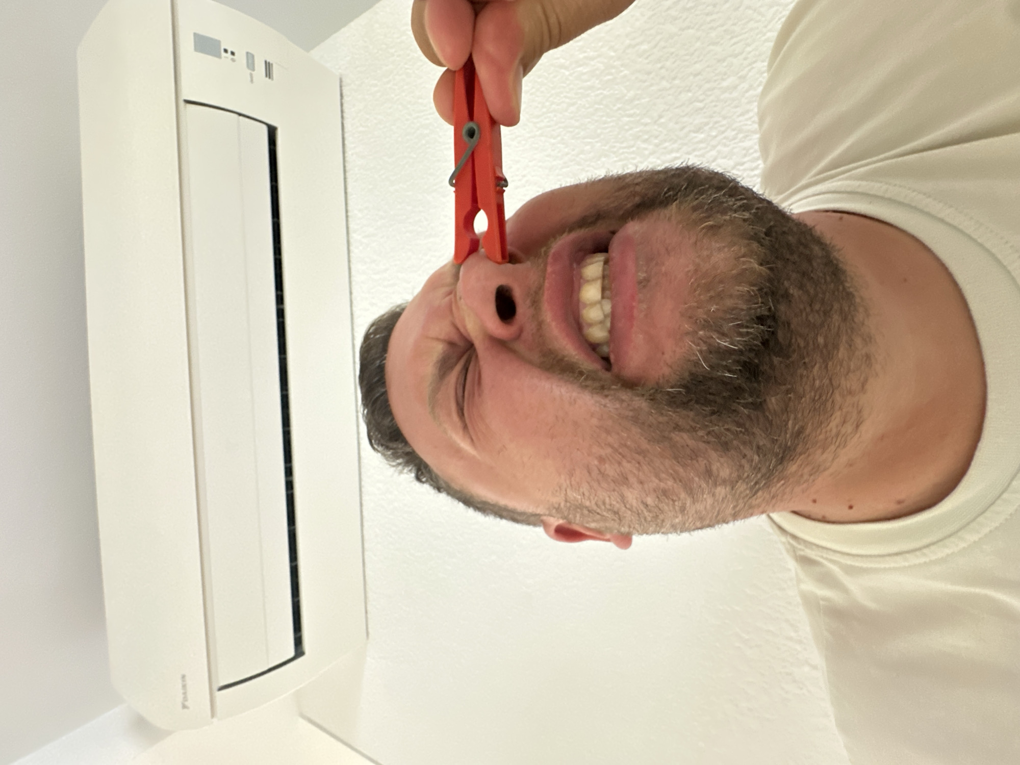 A man with a beard and a white t-shirt is shown from the chest up, with a clothes peg pinching his nose. He is looking upwards, with his eyes closed and his mouth open, revealing his teeth. Above him, a white air conditioning unit is mounted on a white wall. The image was taken in Sant Feliu de Guíxols, Spain.