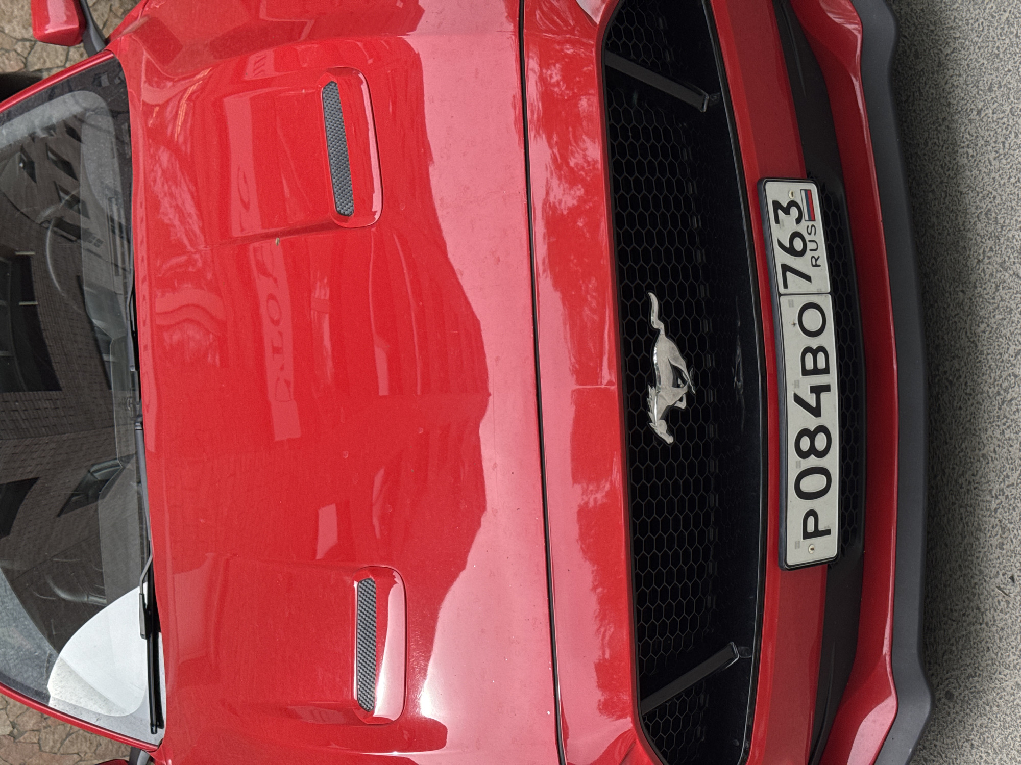 A close-up, vertical shot of the hood and grill of a red Ford Mustang with a Russian license plate, P084BO 763. The car is parked on what appears to be a city street in an unknown location. The focus is primarily on the car's front end, showcasing the hood vents, Mustang emblem, and grill design.