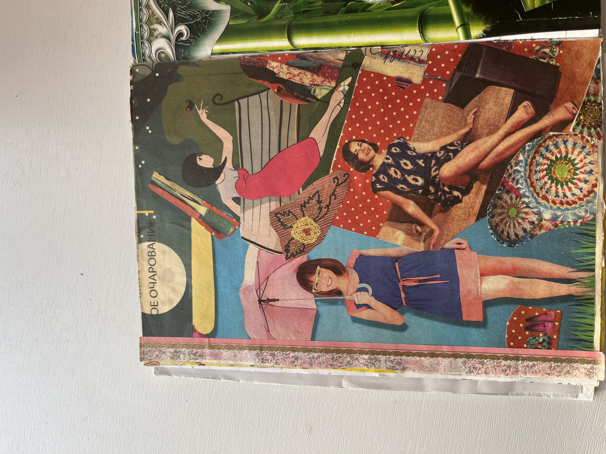 The image is a close-up, overhead shot of a collage on a white wooden surface. The collage features several cutouts of vintage magazine imagery, arranged in a playful, layered composition. On the left side, a woman with short brown hair and glasses stands holding a pink umbrella. She wears a blue dress with a pink belt and trim. To her right, a woman in a red dress is depicted sitting on a bench, reaching out a hand towards a small bird. She has dark bobbed hair. Below this scene, another woman is shown reclining on a patterned armchair, wearing a dark blue dress with a floral pattern. Various decorative elements, such as circular patterned cutouts and fabric swatches, are incorporated throughout the collage. To the right of the main collage, a portion of another collage is visible, featuring green bamboo stalks. The overall aesthetic suggests a retro, artistic theme. The location is not discernible from the image.