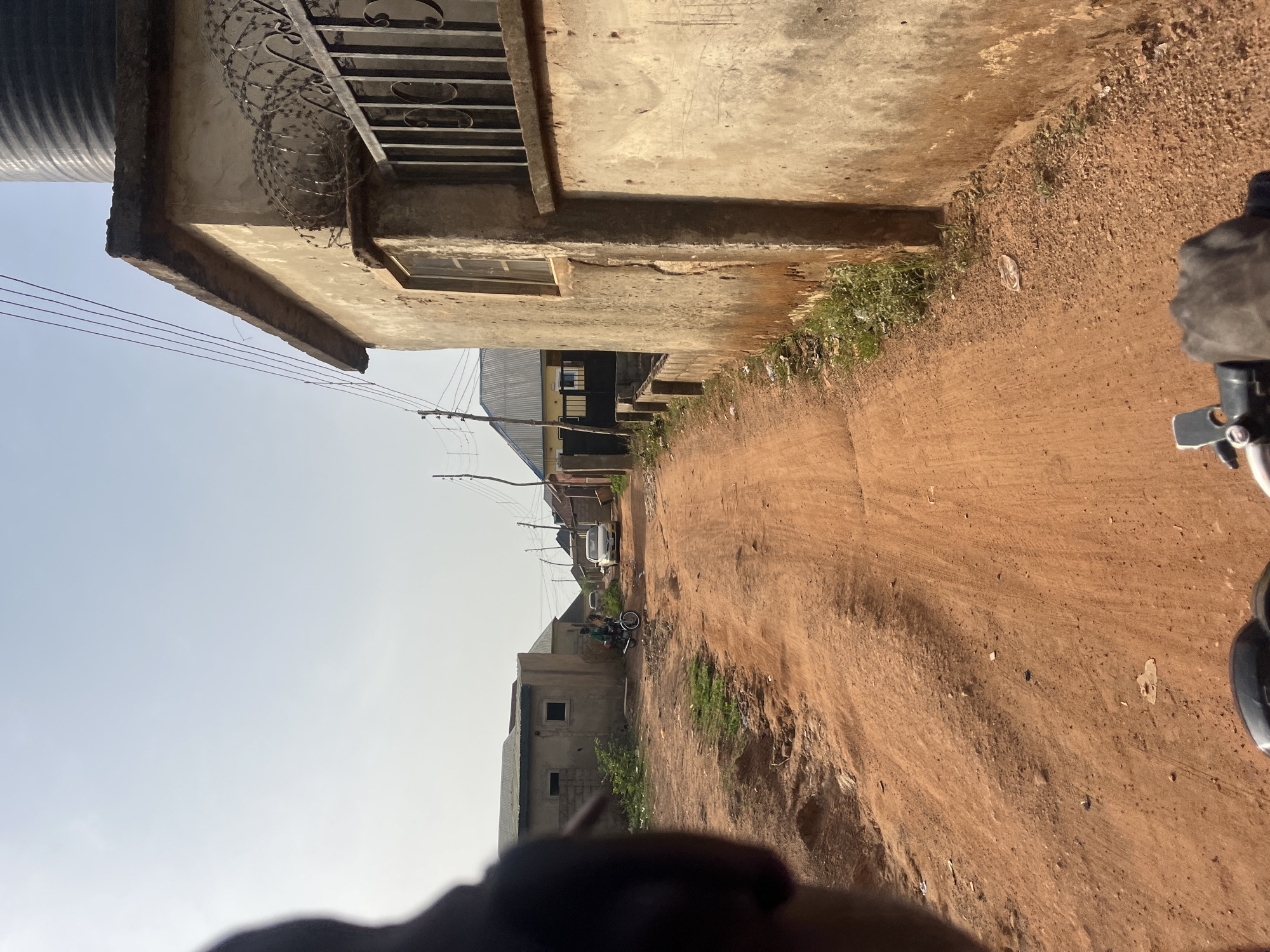 This image captures a daytime scene along an unpaved, reddish-brown dirt road in what appears to be a residential or semi-urban area, consistent with the given location of Abuja, Nigeria. The bright, clear sky indicates sunny weather.
In the foreground on the left, a building with textured, light-colored walls stands, featuring a rectangular window. Above this, a structure with horizontal metal bars and a coil of barbed wire is visible, suggesting security measures. A large, dark cylindrical object, possibly a water tank, is partially seen at the top left edge. Utility lines stretch across the sky from poles visible further down the road.
The dirt road, which takes up a significant portion of the image, shows tire tracks and is bordered by patches of green grass and weeds. Further along the road, more buildings with varying rooflines, including some with corrugated metal, line the sides. A white car is parked in the distance, and closer to the foreground, a motorcycle with at least one individual seated on it is visible, likely an "okada" as commonly found in Nigeria.
The perspective of the photograph suggests it was taken from a vehicle, possibly another motorcycle, given the dark, blurry forms visible in the bottom right and bottom left corners, resembling parts of handlebars or a person's body. The overall scene depicts a typical street environment in a developing locale. No discernible text is visible in the image.