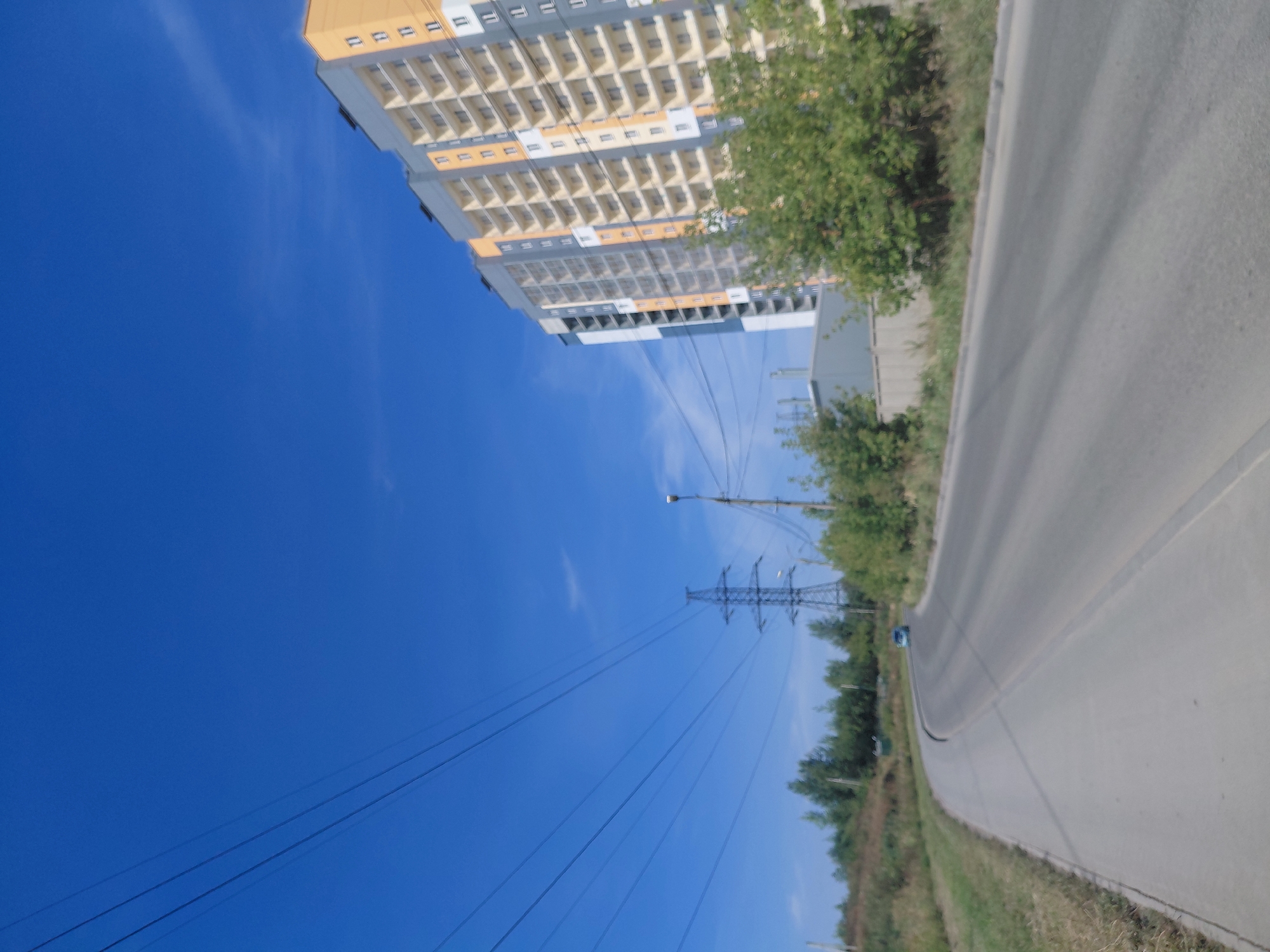 The image shows a street in Novosibirsk, Russia, on a clear day. A modern, multi-story apartment building with yellow and gray facades is visible on the right side of the frame. In the distance, a power line tower stands against the blue sky. A road curves through the scene, with a blue car driving on it in the middle ground. Trees and vegetation line the sides of the road, and a sidewalk is present on the left. The sky is a vibrant blue with a few wispy clouds.