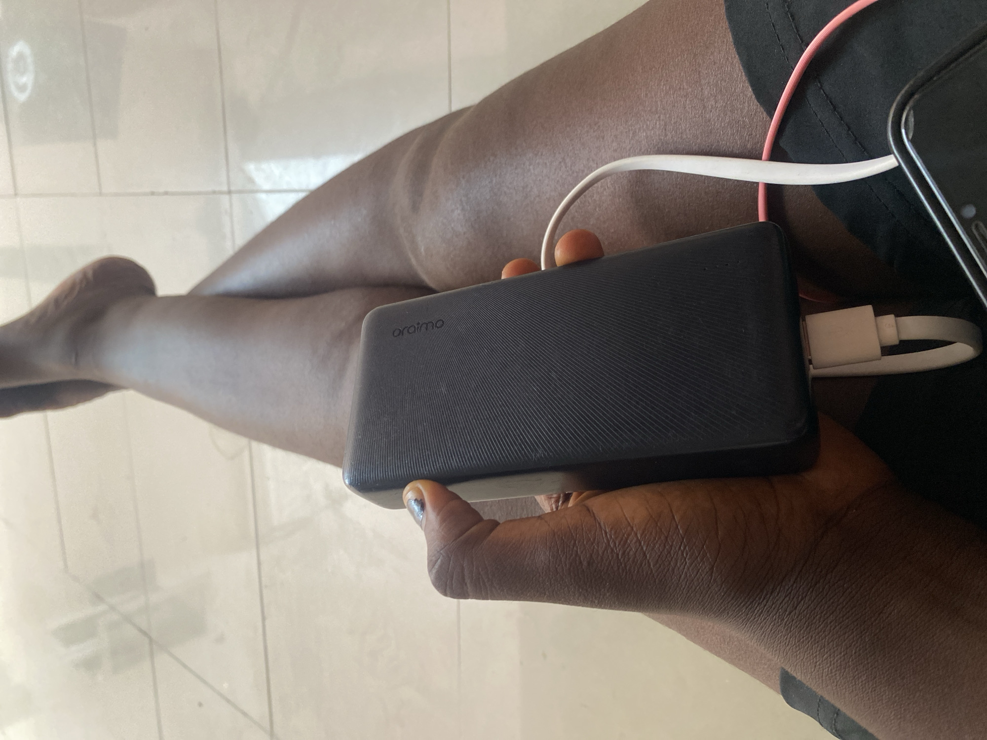 A close-up, overhead shot shows a person's legs and hand, holding a black power bank. The power bank is connected to a white charging cable, which is plugged into a smartphone not fully visible in the frame. The person's hand has dark skin, with a bit of nail polish on one finger. The background appears to be a tiled floor. The person is wearing black shorts. The scene is likely indoors, possibly in a home in Bulamare, Nigeria.