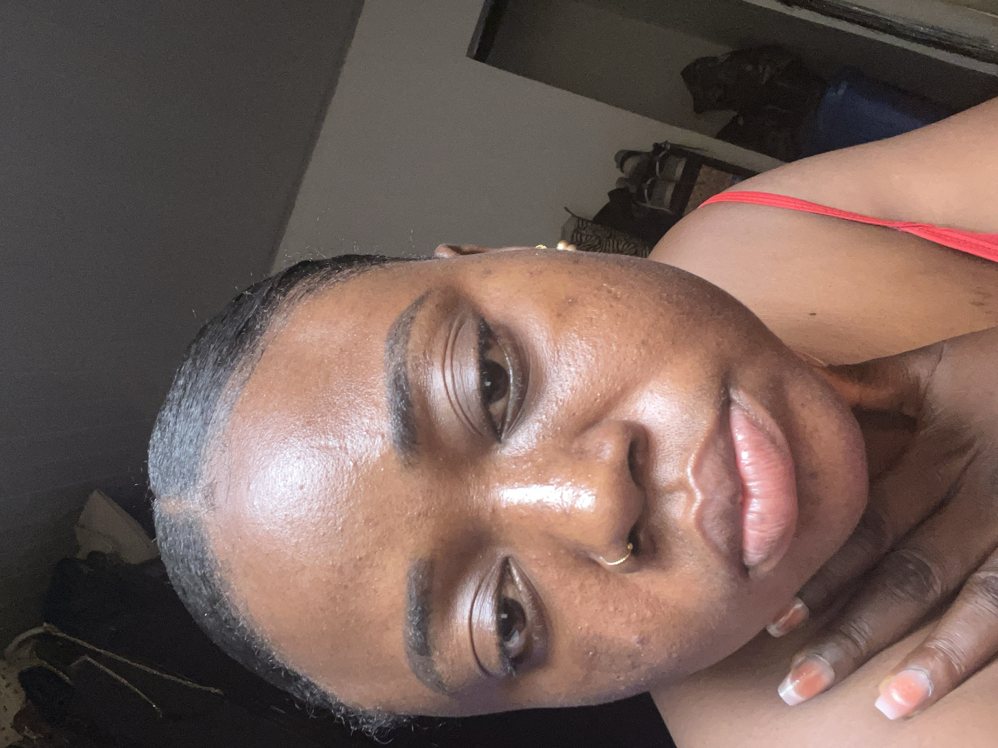 A close-up shot of a woman's face fills the frame. She has dark skin, dark eyebrows, and brown eyes. A small gold nose ring is visible. Her lips appear full and pink. Her right hand is near her face, with fingernails that are painted with a light design. A red strap of clothing is visible on her upper right side. The background is blurred and out of focus, suggesting an indoor setting. The location of Abuja, Nigeria, cannot be verified from this image.