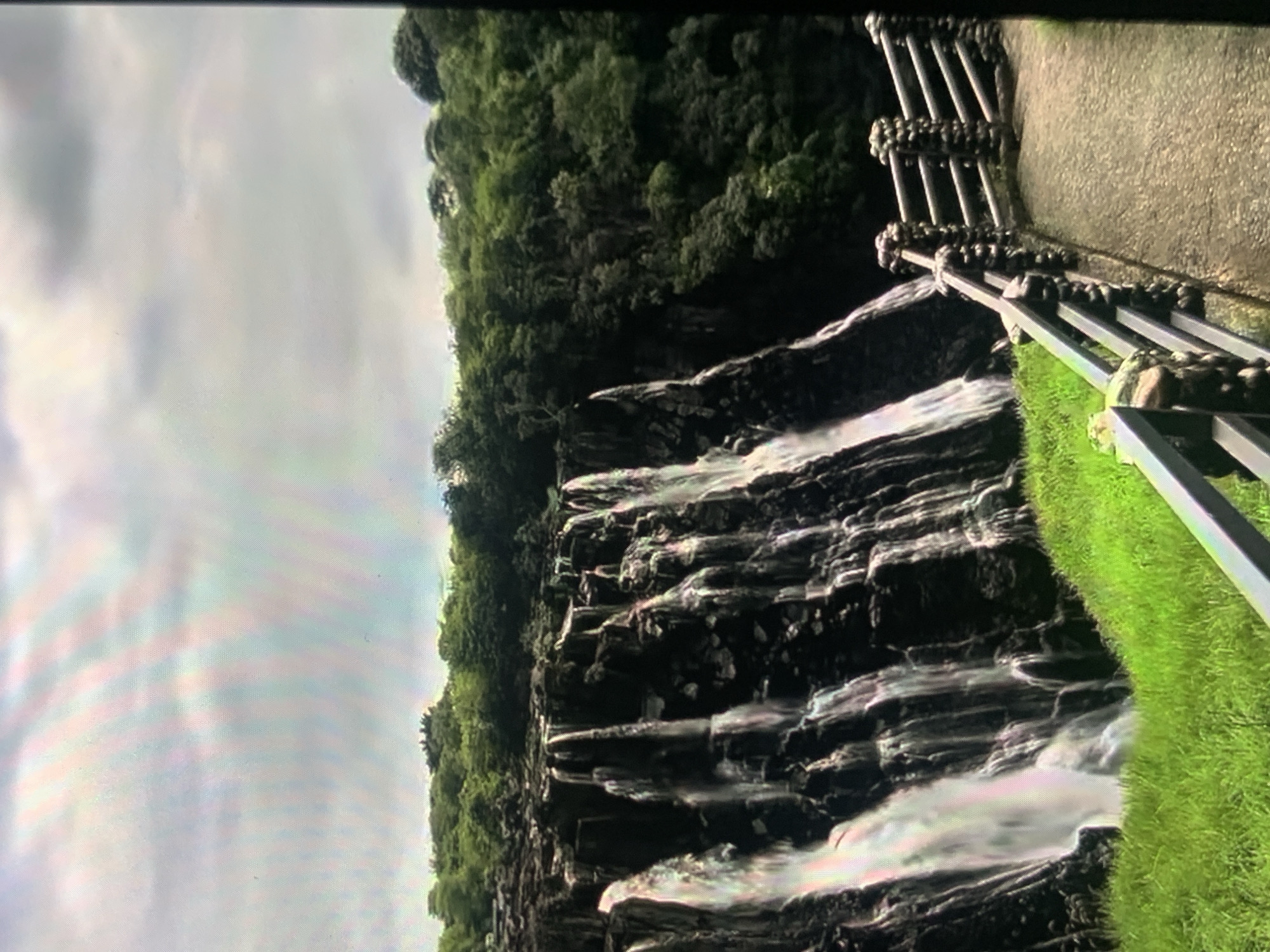 The image depicts a wide, multi-tiered waterfall cascading down dark, rocky cliffs. Lush green vegetation, including trees and grass, surrounds the waterfall. A paved walkway with a stone and metal railing follows the edge of the cliff, offering a viewpoint. The sky above is overcast with gray clouds. This scene is not located in Stavropol, Russia.