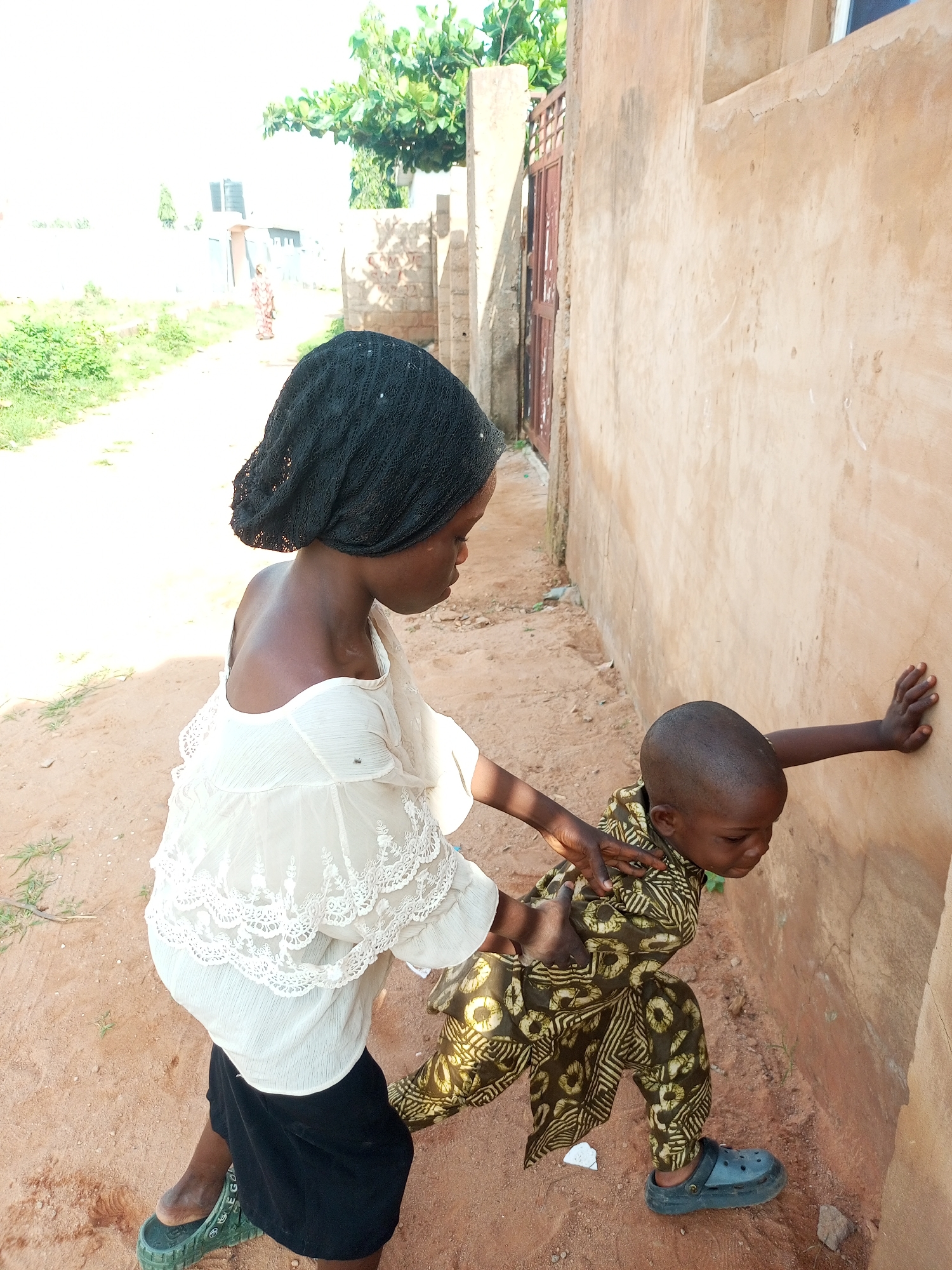 In Tempure, Nigeria, a young girl wearing a black headscarf and a white lacy blouse, with a black skirt, is shown interacting with a younger child. The younger child, in a patterned garment, is leaning against a tan-colored wall. The girl has her hands on the younger child's back. The environment appears to be a dirt path next to the wall, with some vegetation visible in the background.