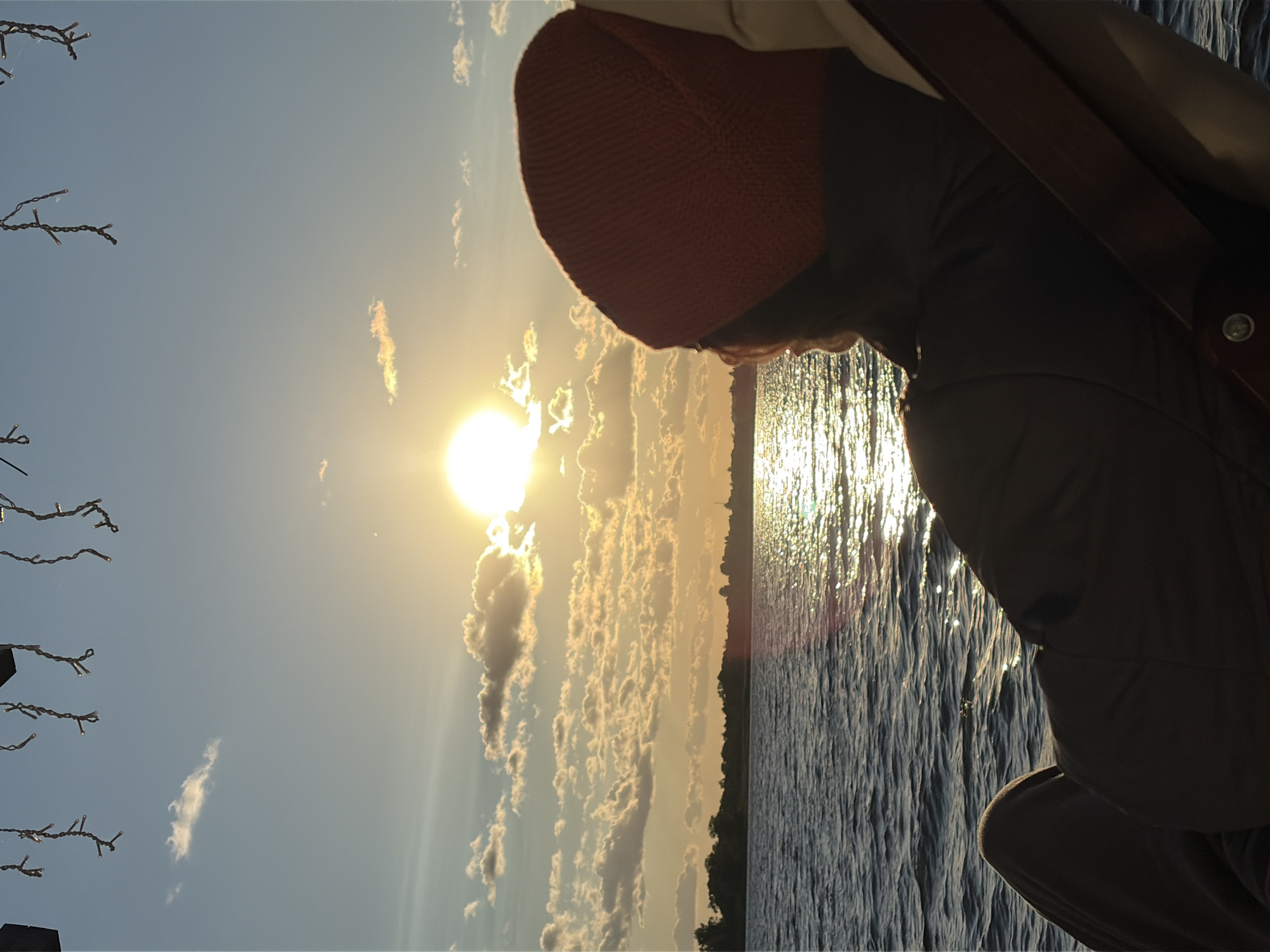 An over-the-shoulder shot shows a person wearing an orange beanie and a dark jacket, sitting in a lounge chair and looking out at a sunset over a body of water. The sun is low in the sky, casting a bright reflection on the water. Clouds are scattered across the sky, some tinged with the orange and yellow hues of the sunset. A dark, tree-lined shore is visible in the distance. Overhead, string lights are partially visible against the blue sky. The scene appears to be set on a calm evening in Vidogoshchi, Russia.