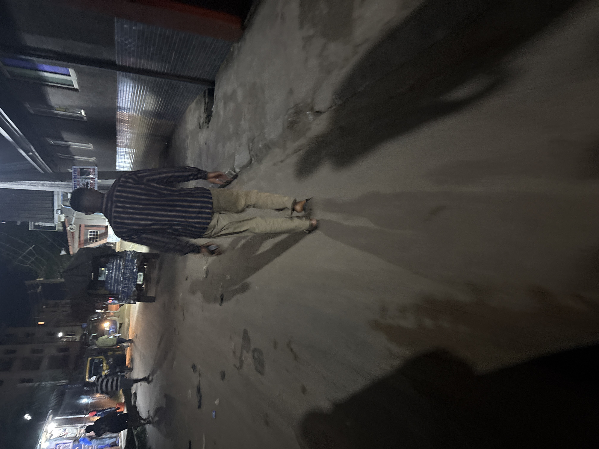 The image depicts a nighttime street scene in Onitsha, Nigeria. A man, seen from behind, is walking away from the camera down a paved street. He is wearing a vertically striped long-sleeved shirt and light-colored trousers. In the background, to the left, are buildings with lights on, and several people are visible, some walking and others standing. A blue three-wheeled vehicle, likely a commercial taxi, is parked on the street. To the right of the street, there is a building with a dark facade and several windows. The street surface appears rough and uneven, with some debris scattered on it. Long shadows are cast across the ground, indicating the presence of artificial lighting. The overall atmosphere is that of a busy urban environment at night.