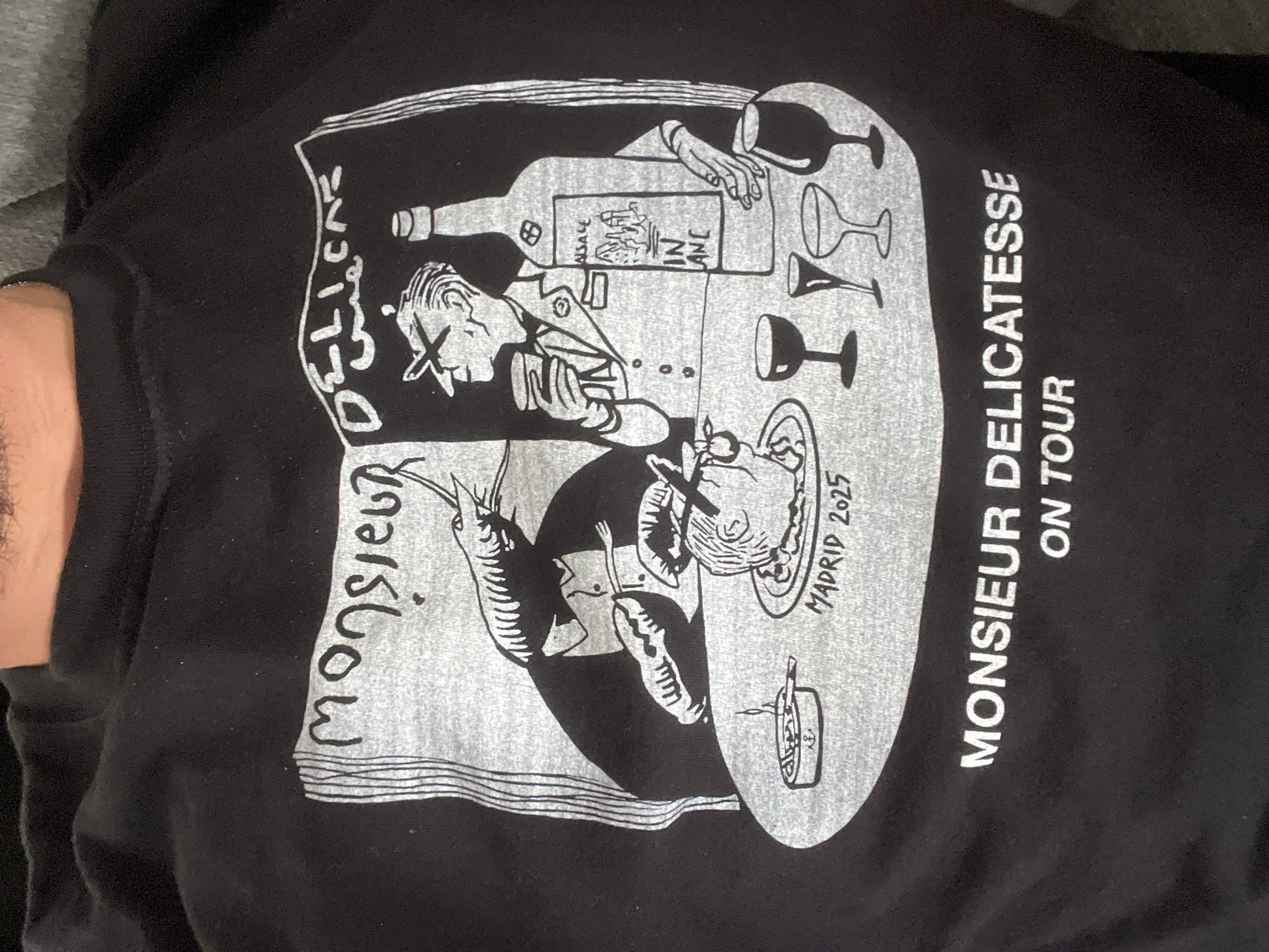 The image displays a black t-shirt featuring a detailed, cartoon-style graphic in white and light gray, possibly being worn by someone, with a portion of an arm visible on the left. The scene depicts a culinary or wine-tasting event.
**Subjects:** The graphic features two stylized male figures. A larger figure, dressed in what appears to be a uniform or suit, holds a glass and gestures towards the setting, possibly presenting or serving. Beside him, a large wine bottle labeled "ALSACE" and "VIN BLANC" is prominent, surrounded by various empty and filled wine glasses. Below, a smaller figure is seated at a table, appearing to eat with a fork near his mouth. The table is laden with food items, including a plate with what looks like seafood, a large shrimp or prawn, a small bowl with an anchor motif, and another small dish.
**Activity or event taking place:** The figures are engaged in dining and drinking, suggesting a gourmet experience, wine tasting, or a celebratory meal. The "ON TOUR" text implies a series of events.
**Notable details:** The artwork has a distinct hand-drawn, illustrative quality, using a limited color palette of black and white/gray. The text "MADRID 2025" is visible near the smaller figure, indicating a specific future event location and year. No specific time of day, weather, or discernible emotions are depicted.
**Visible text:** Key phrases include "MONSIEUR DELICATESSE" (written vertically on the left side of the graphic and on the right side, followed by "ON TOUR"), "ALSACE" and "VIN BLANC" on the wine bottle label, and "MADRID 2025."