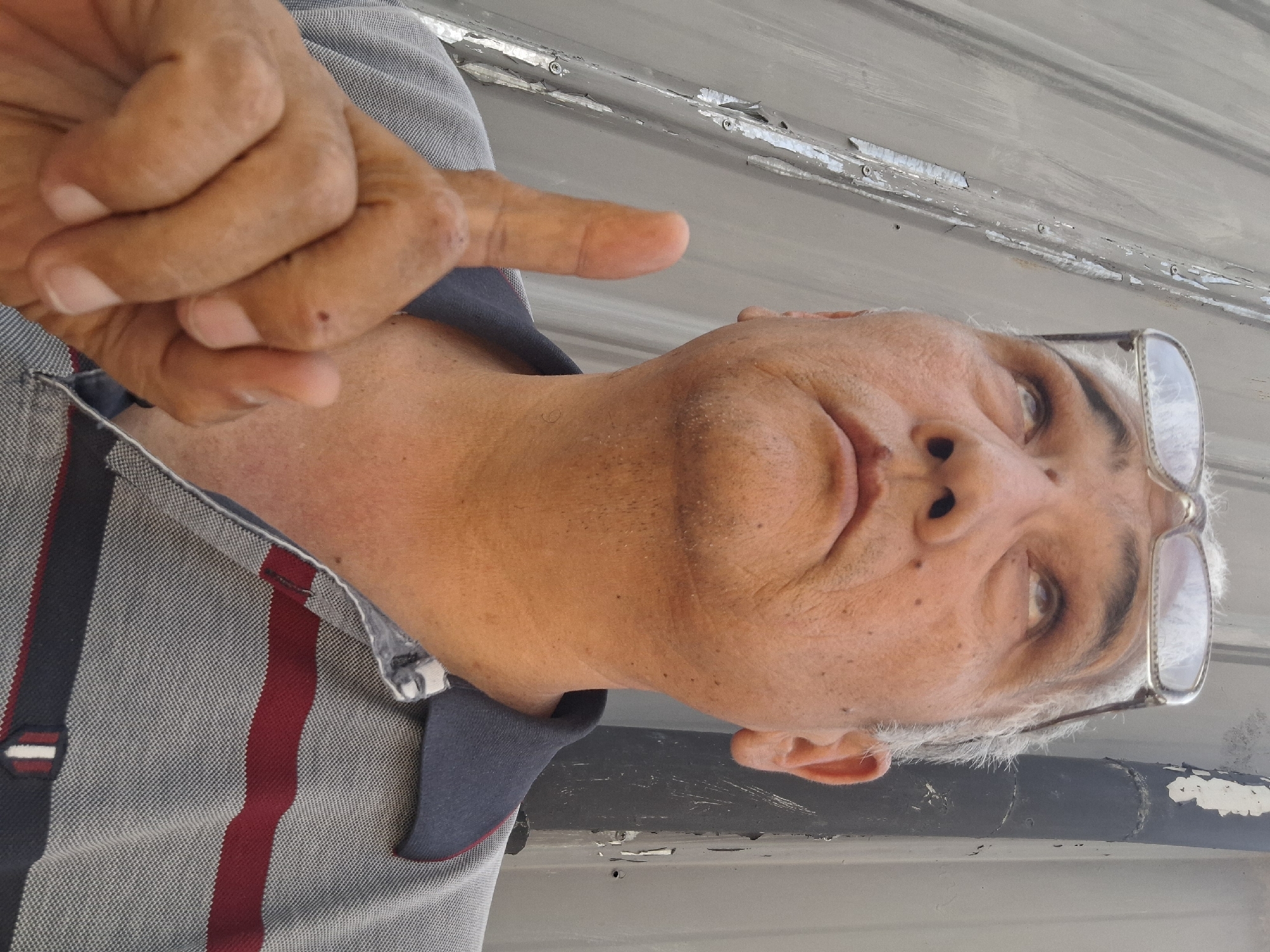 A close-up, low-angle selfie shows an older East Asian man with graying hair and glasses looking upwards. His right index finger is raised. He is wearing a gray polo shirt with red and dark blue stripes. The background is a corrugated metal wall, and a black pipe with peeling paint is visible to the right. The image appears to have been taken outdoors during the day. There is no discernible information in the image to suggest the location is Almaty, Kazakhstan.
