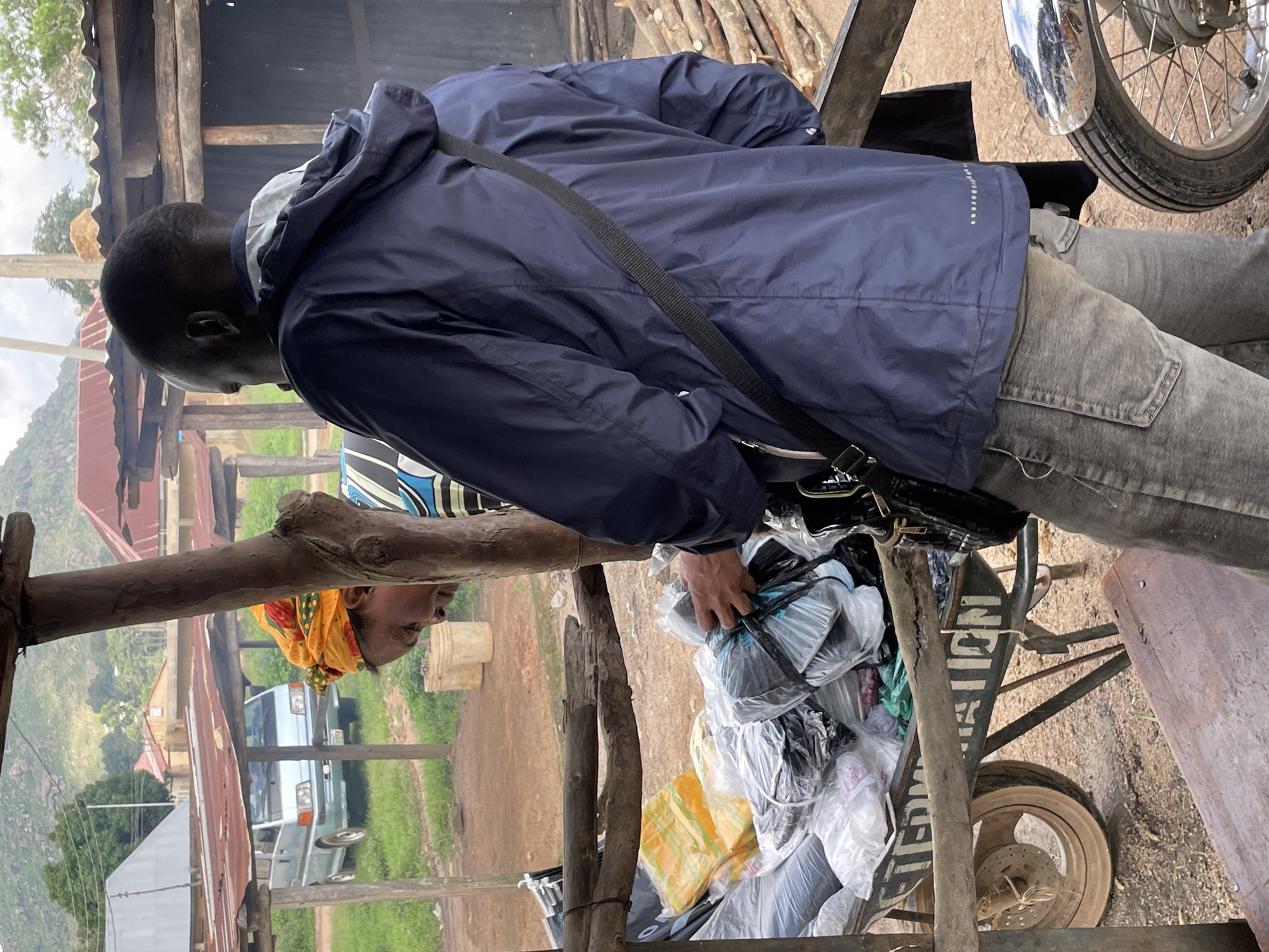 In Wang, Nigeria, two individuals are present. In the foreground, a person wearing a dark blue jacket and grey jeans is reaching towards a cart filled with various items, many of which are packaged in plastic bags. The cart appears to be a wheelbarrow with wooden supports and a visible wheel with spokes. Behind this individual, another person, partially obscured, is wearing a yellow headscarf and a striped top. They are looking down at the cart's contents. The background reveals a rural or semi-rural setting with a corrugated metal roof structure, a light blue van, and vegetation. The ground is a mix of dirt and scattered debris. The overall scene depicts a transaction or an exchange taking place at an outdoor market or roadside stall.