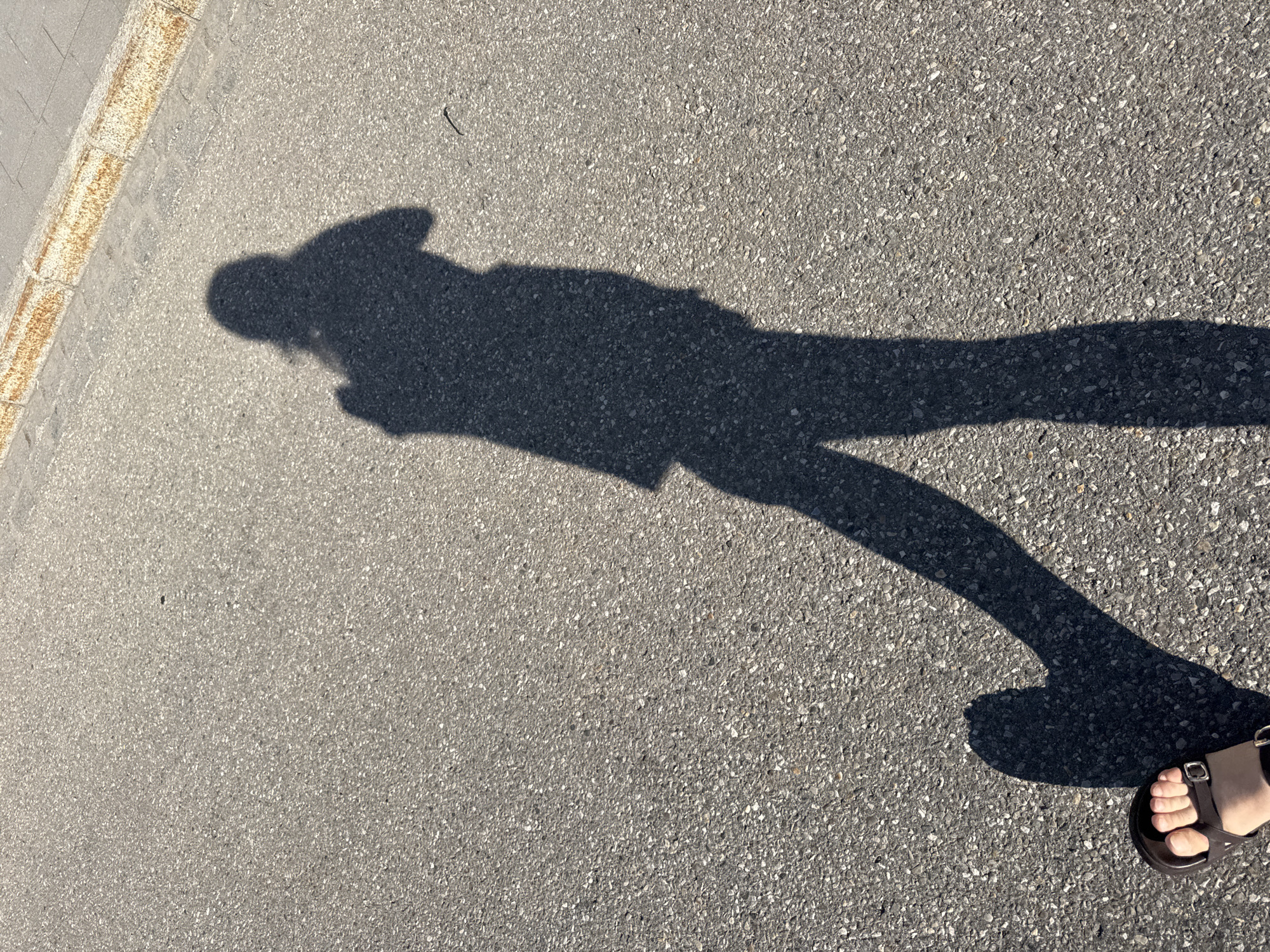 The image shows the shadow of a person on a paved surface, likely a street or sidewalk, in Höhenkirchen-Siegertsbrunn, Germany. The shadow is cast by the sun and depicts a person wearing a shirt and possibly pants, with a shadow of a hand holding a device visible near the head. A foot wearing a sandal is also visible in the lower left corner of the image, casting its own shadow. The paved surface appears to be asphalt, with some gravelly texture. A curb with a rusty appearance is visible along the top right edge of the image. The lighting suggests it is daytime.
