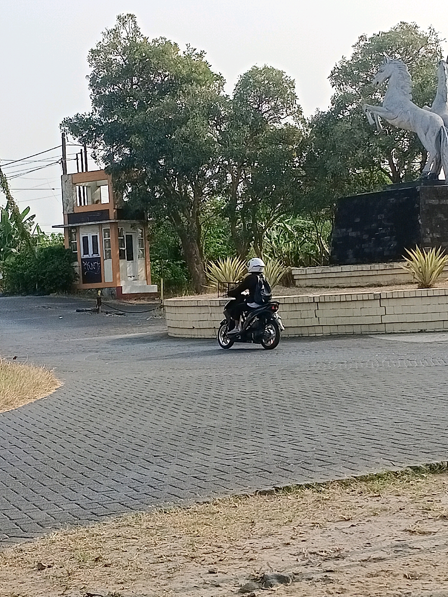 The image shows a person riding a dark-colored scooter on a paved road in Tawangsari, Indonesia. The rider is wearing a white helmet and a dark jacket, and a backpack is visible on their back. The scooter is moving from left to right across the frame. In the background, there is a small, multi-level structure with windows and doors, appearing to be some sort of kiosk or guardhouse, adorned with graffiti. Behind this structure are lush green trees and a statue of two horses. The road is paved with interlocking bricks, and dry grass and dirt line the sides of the road. The sky is overcast.