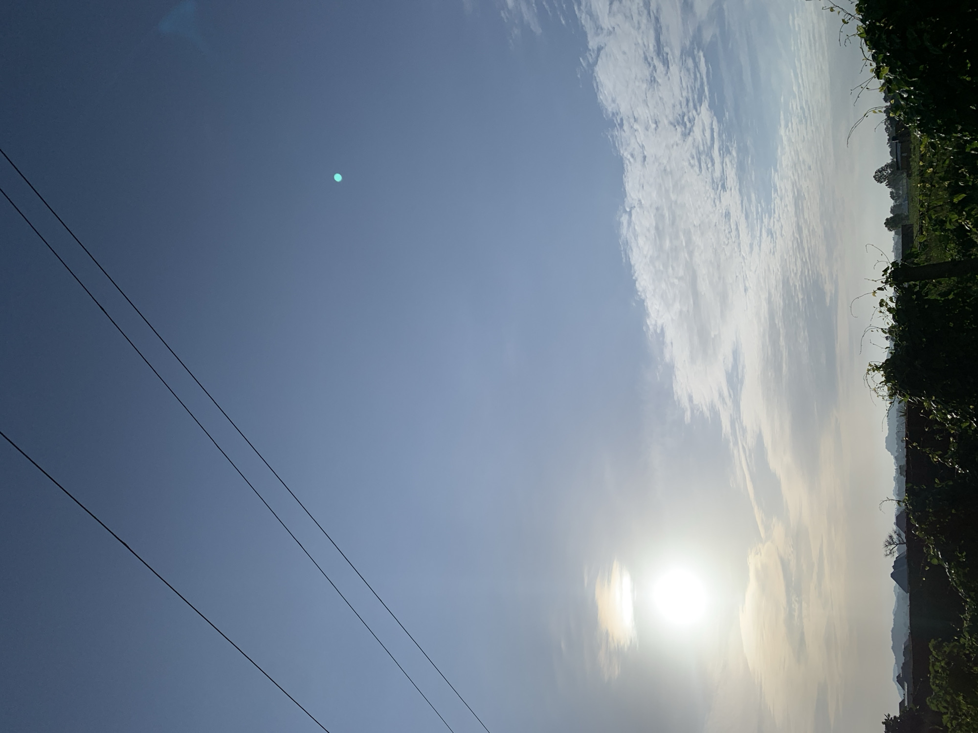 The image depicts a wide shot of the sky with a sliver of land and foliage in the foreground, suggesting an outdoor environment, potentially in Kuru Karama, Nigeria. Three dark, parallel power lines stretch diagonally across the upper portion of the frame, against a clear blue sky. Toward the upper right, a small, bright cyan circular anomaly is visible, possibly a lens flare or artifact. The sun, a bright white orb, is positioned in the lower left quadrant, partially obscured by thin, wispy clouds that streak horizontally. These clouds extend across the middle of the frame, with more dense, textured white clouds appearing in the middle right. The horizon line is low, showcasing a silhouette of distant, rolling hills or mountains against the bright sky. In the immediate foreground, dark green foliage, appearing to be vines or bushes, occupies the bottom portion of the image, with some organic shapes and darker vertical elements, likely posts or thicker stems, interspersed. The overall scene suggests a landscape under a bright sky, with man-made infrastructure (power lines) present.