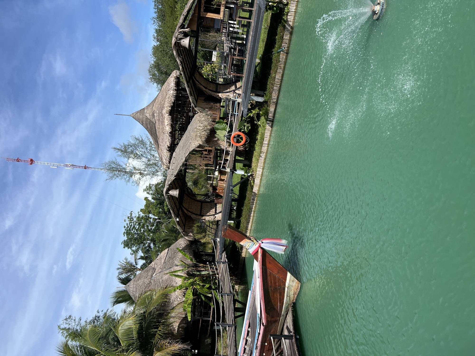 The image depicts a picturesque scene in Tambon Pa Kae Bo Hin, Thailand, featuring a tranquil turquoise body of water. In the foreground, a traditional longtail boat, painted a deep red, is moored to a wooden dock. The boat is adorned with a colorful fabric tied to its bow. Across the water, a series of thatched-roof huts are situated on elevated wooden platforms, accessible by walkways. These structures appear to be open-air dining or seating areas, with tables and chairs visible within some of them. One of the huts has a bright orange life preserver hanging on its side. A small fountain in the water sprays water upwards, creating a pleasant visual effect. Palm trees and lush greenery surround the huts, adding to the tropical ambiance. In the background, a tall communication tower rises against a bright blue sky dotted with wispy clouds. The scene suggests a peaceful, resort-like setting, ideal for relaxation and enjoying the natural beauty of the region.