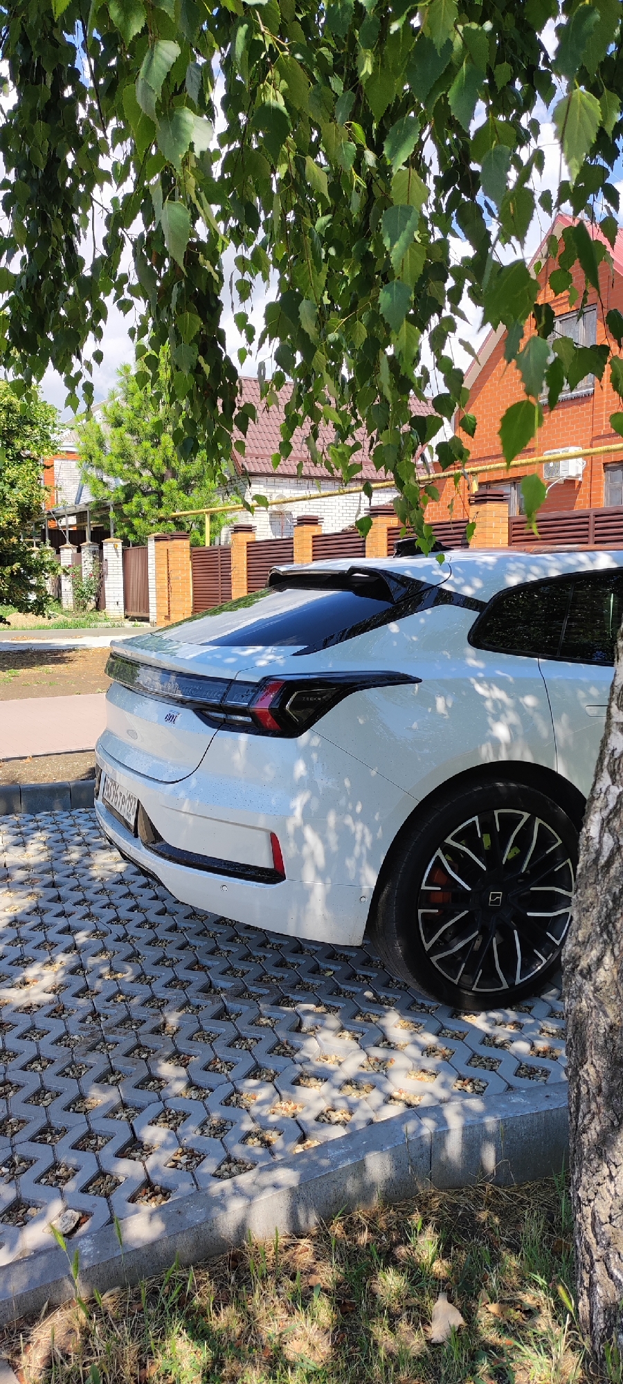 A white car is parked on a gravel-filled interlocking concrete paver driveway, viewed from the rear quarter panel. The car, a Zeekr 001, has a sleek design with distinctive taillights and a prominent rear spoiler. The wheels are black with silver accents. A tree trunk partially obstructs the right side of the image, and branches with green leaves hang over the scene, casting dappled shadows.
In the background, there are residential buildings with brick walls and tiled roofs, characteristic of rural or suburban Russia. A wooden fence separates the property from the street. The sky is visible through the leaves, suggesting it is daytime with some clouds. The overall impression is of a quiet, sunny day in a residential area, likely in Dinskaya, Russia. No people are visible in the image. There is visible text on the car's license plate, which appears to be "Р976ТР 197".