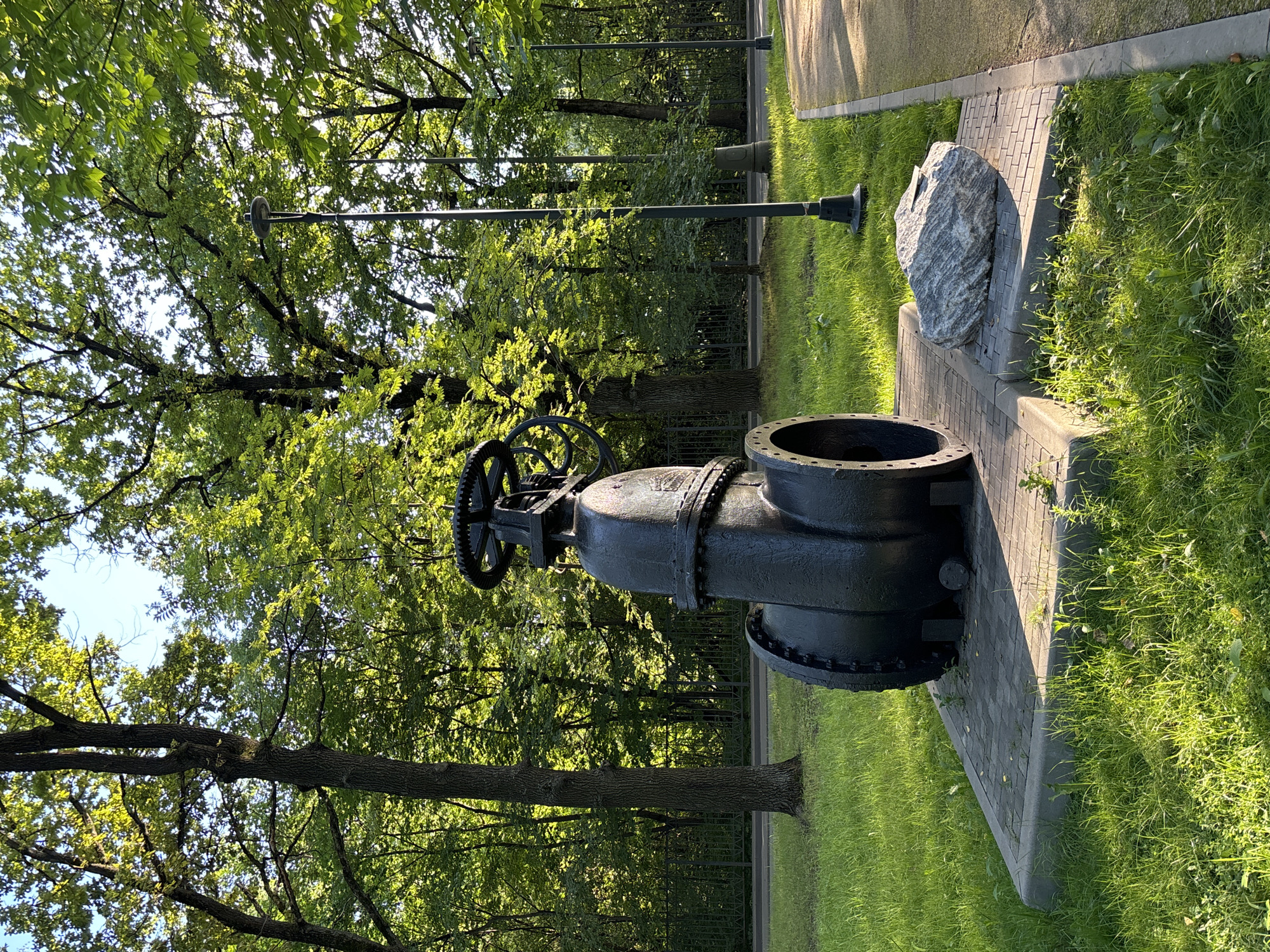 This is a photograph of a large, black, industrial valve displayed outdoors on a paved pedestal in a park. The valve is positioned on the left side of the image and is the primary subject. It appears to be made of metal and has a large wheel at the top for operation. Adjacent to the pedestal, on the right, is a large, grey, irregular-shaped rock. The background consists of lush green grass, trees with vibrant green foliage, and a clear blue sky. A paved pathway runs along the right edge of the frame, and several lampposts are visible in the mid-ground. The scene suggests a public park setting, possibly in Moscow, Russia, given the style of the lampposts and the mature trees. There are no people or animals visible in the image. The lighting indicates it is daytime, with sunlight casting shadows on the grass and the valve.