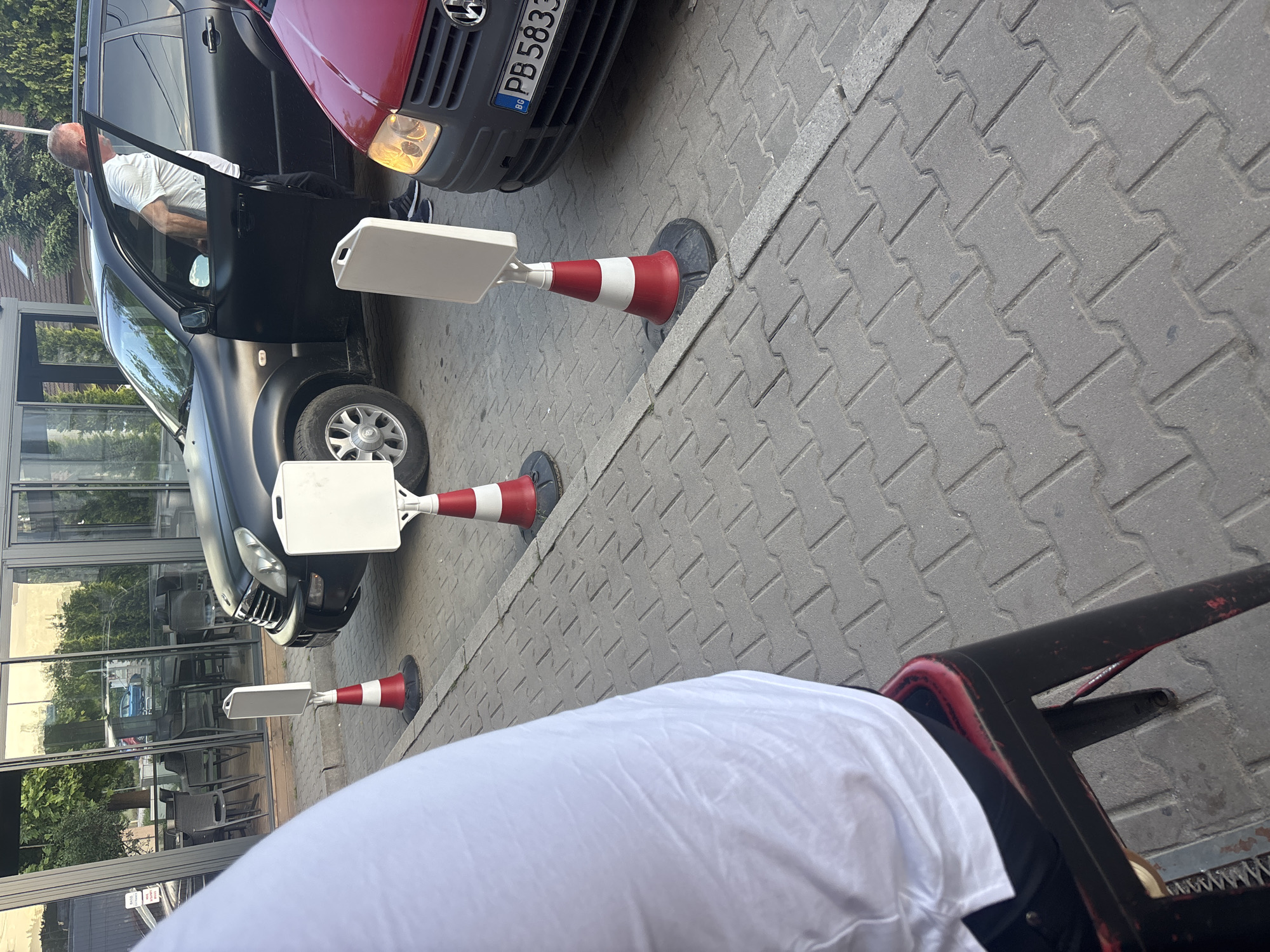 A medium-angle view shows a partially occupied black SUV, its door open to reveal a man in a white shirt exiting, parked on a brick-paved street in Plovdiv, Bulgaria. In front of the vehicle are two parking cones, red and white striped, each topped with a small, flat, white square sign. A red car is partially visible in front of the SUV; its license plate reads "PB 5833 BG". A building with glass windows reflecting exterior trees and furniture is behind the parked vehicles, suggesting a restaurant or cafe. The lower part of a person's white shirt and the corner of a black metal chair are visible in the foreground, suggesting the photo was taken from a seated position.