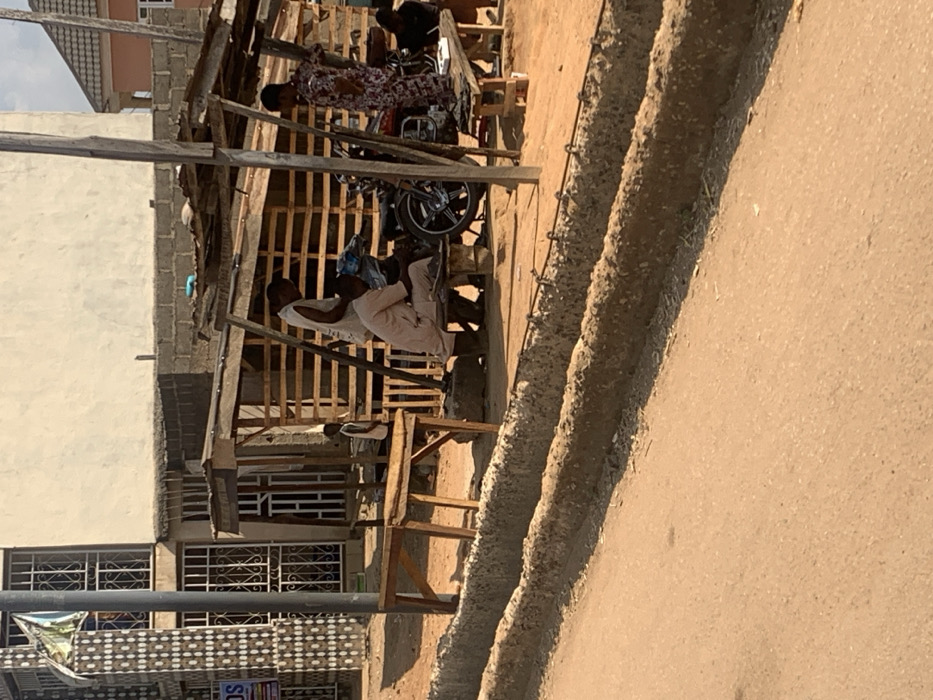 This image captures an outdoor street scene in Egbedi, Nigeria, under daylight. In the foreground, a reddish-brown dirt road is present, bordered on the right by two parallel, unfinished concrete structures, possibly for drainage, which feature exposed rebar.
Midground features a makeshift wooden shelter with a corrugated roof, supported by poles. Beneath this shelter, a dark-colored motorcycle is centrally positioned. A man in light-colored traditional attire is seated on a low wooden bench in front of the motorcycle, appearing to be engaged with it. Another man, wearing a white sleeveless top, stands directly behind the seated man. To their right, a woman in a multi-colored patterned dress stands, holding an object in her right hand. A fourth individual is partially visible, seated on a bench to the far right. Further left, adjacent to the shelter, a child in a white vest is partially visible behind an empty wooden table.
The background on the left features a multi-story building with a light-colored facade, distinguished by a pillar with decorative dark and light-colored tiles and multiple windows fitted with metal security grates. Other less defined residential or commercial buildings are visible further in the background.