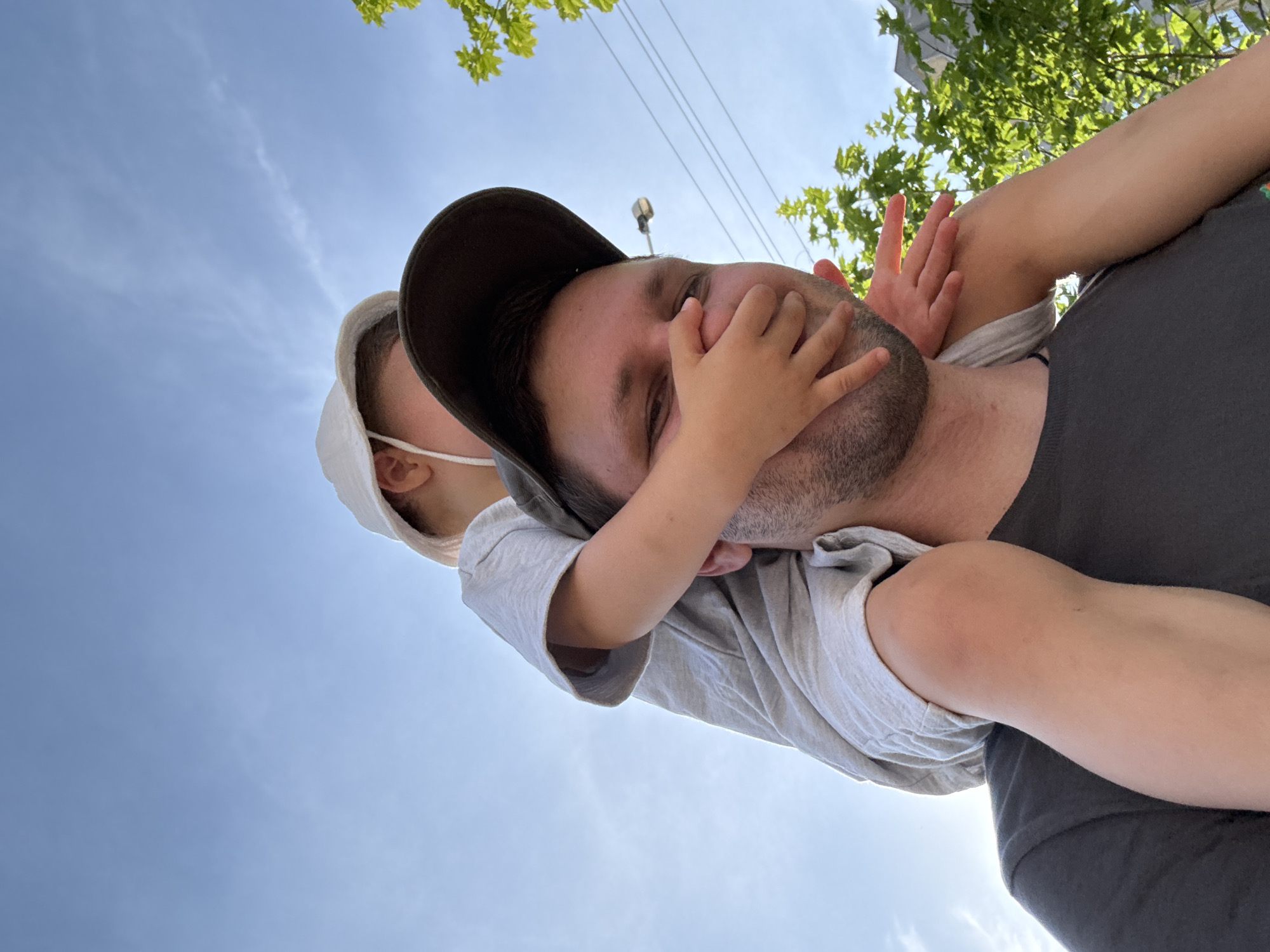 The image shows a low-angle, close-up shot of a man and a child against a bright blue sky with some clouds. The man is wearing a dark baseball cap and a grey t-shirt, and he has a beard. The child, wearing a white sun hat and a white face mask, is perched on the man's shoulders and has their hands over the man's mouth. The child's hands are small and appear to be playfully covering the man's mouth. The man's eyes are visible, and his expression is difficult to discern due to the child's action, but there's a sense of interaction and lightheartedness.
The background is dominated by a clear sky, suggesting it is daytime. There are some green leaves from a tree visible in the upper right corner, and a few power lines also appear in the sky. The perspective suggests the photo was taken from below, looking upwards. The sunlight is bright, casting a gentle glow. The overall mood is one of playful intimacy and connection between the man and the child. No text is visible in the image.