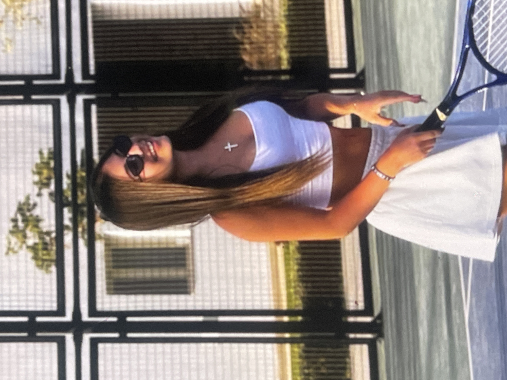 An outdoor, eye-level, full-shot photograph captures a young woman posing on what appears to be a tennis court in Poltava, Ukraine, during daylight hours under sunny conditions. The woman, with long brown hair, is shown from the waist up, smiling broadly while looking towards the viewer. She is wearing dark sunglasses, a white sleeveless crop top, and a white tennis skirt. Around her neck, she wears a silver cross pendant. On her left wrist, she has multiple bracelets. She holds a blue and black tennis racket with both hands, positioned in front of her.
The background features a grid-like fence or barrier, possibly a chain-link fence or a similar structure, with some blurred greenery visible behind it, suggesting trees or bushes. The court surface appears to be a dark blue or gray color, with faint white lines visible. The overall mood is casual and pleasant, indicated by her relaxed pose and smile. No discernible text is visible in the image.