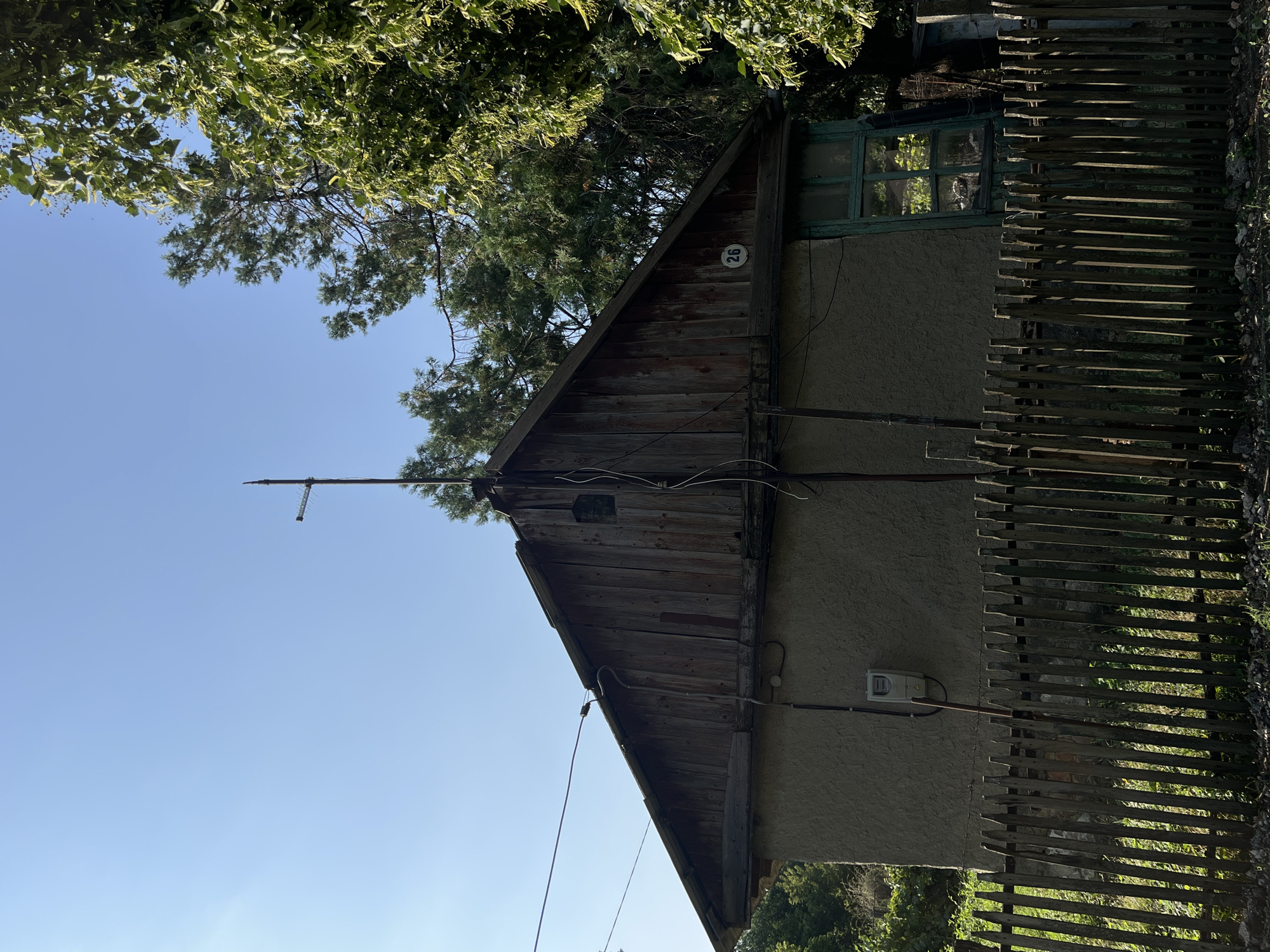 The image depicts the side and front of a single-story house with a weathered wooden gable roof. The house has a rough, stucco-like exterior wall in a neutral gray color. A simple wooden picket fence runs along the front of the property. A green-framed window with multiple panes is visible on the right side of the house. A utility meter is mounted on the exterior wall. Electrical wires are visible running across the roof and down the side of the house, connected to an antenna on the peak of the roof. A circular plaque with the number "26" is affixed to the wooden gable. Lush green trees and foliage surround the house, suggesting a rural or suburban setting. The sky is clear and blue. The scene is likely a residential property in the city of Leova, Moldova.
