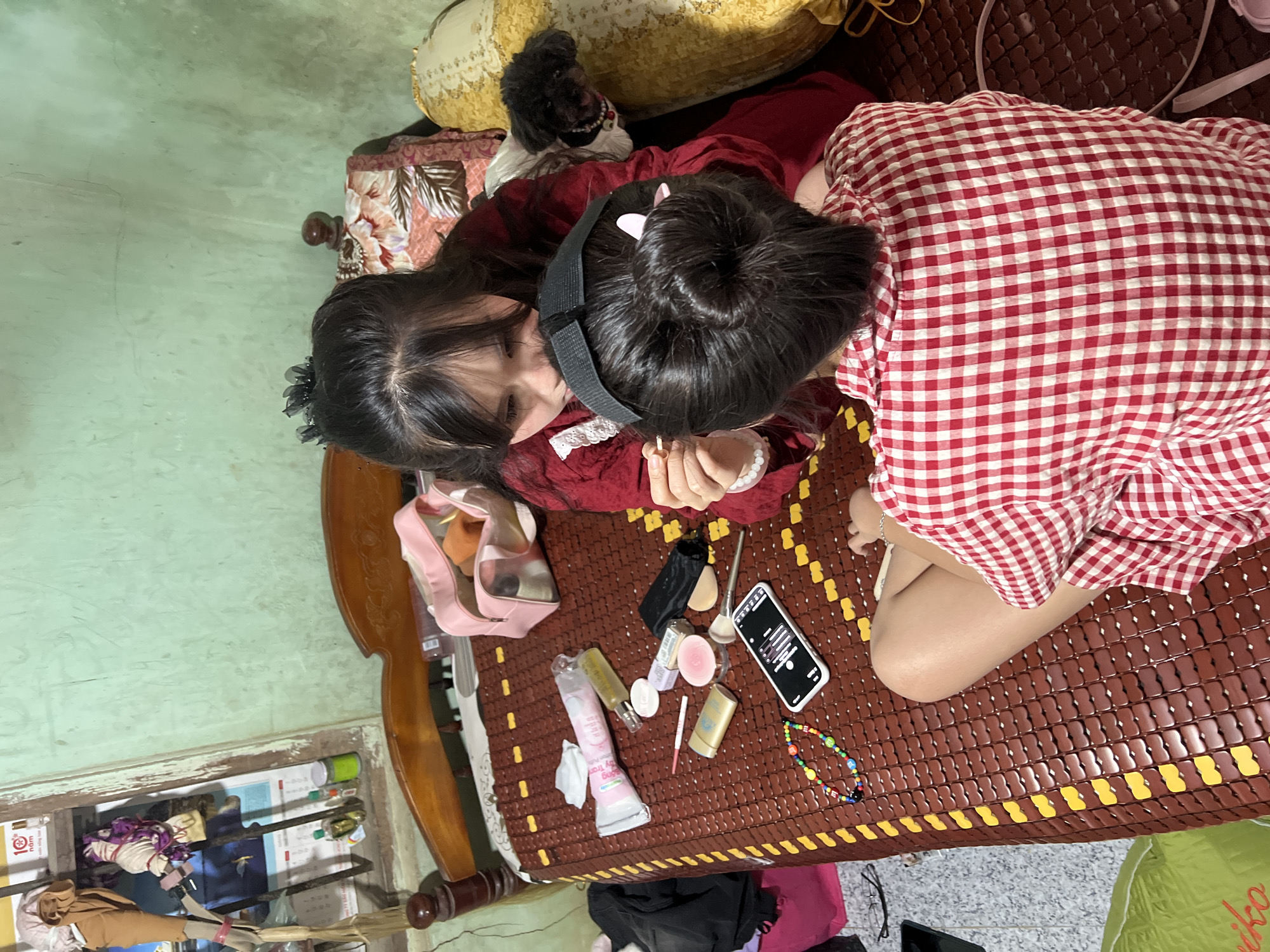 The image presents an overhead view of two young women casually relaxing indoors, likely in a home in Bình An, Vietnam. They are situated on a traditional Vietnamese woven mat (chiếu) characterized by brown and yellow striped patterns.
One woman, positioned on the left side of the frame, has long dark hair and is wearing a deep red long-sleeved top with white lace detailing around the collar. A white pearl bracelet adorns her wrist. She is looking downwards, her face partially obscured by her hair. Behind her head, a decorative pillow and a small black toy dog or doll are visible.
The second woman, to her right, is lying on her stomach, facing away from the camera. Her dark hair is styled into a bun, secured with a black headband, and she is dressed in a red and white checkered short-sleeved top or dress. Her bare legs are visible towards the lower right of the frame.
Between the two women, on the mat, is an assortment of personal items: a pink mesh makeup bag, various makeup products including compact powder, brushes, tubes of cream, a foundation bottle, lipstick, and cotton pads. A black smartphone displaying icons (possibly a music interface) and a colorful beaded necklace are also present.
The surrounding environment indicates an indoor setting, likely a bedroom. A wooden bed frame is partially visible on the left and bottom edges. The floor below the bed appears somewhat cluttered with clothes, bags, bottles, and papers, one of which faintly displays "10 nam" (10 years). The wall in the background is a muted green. The overall scene suggests a relaxed and informal moment between the two individuals.