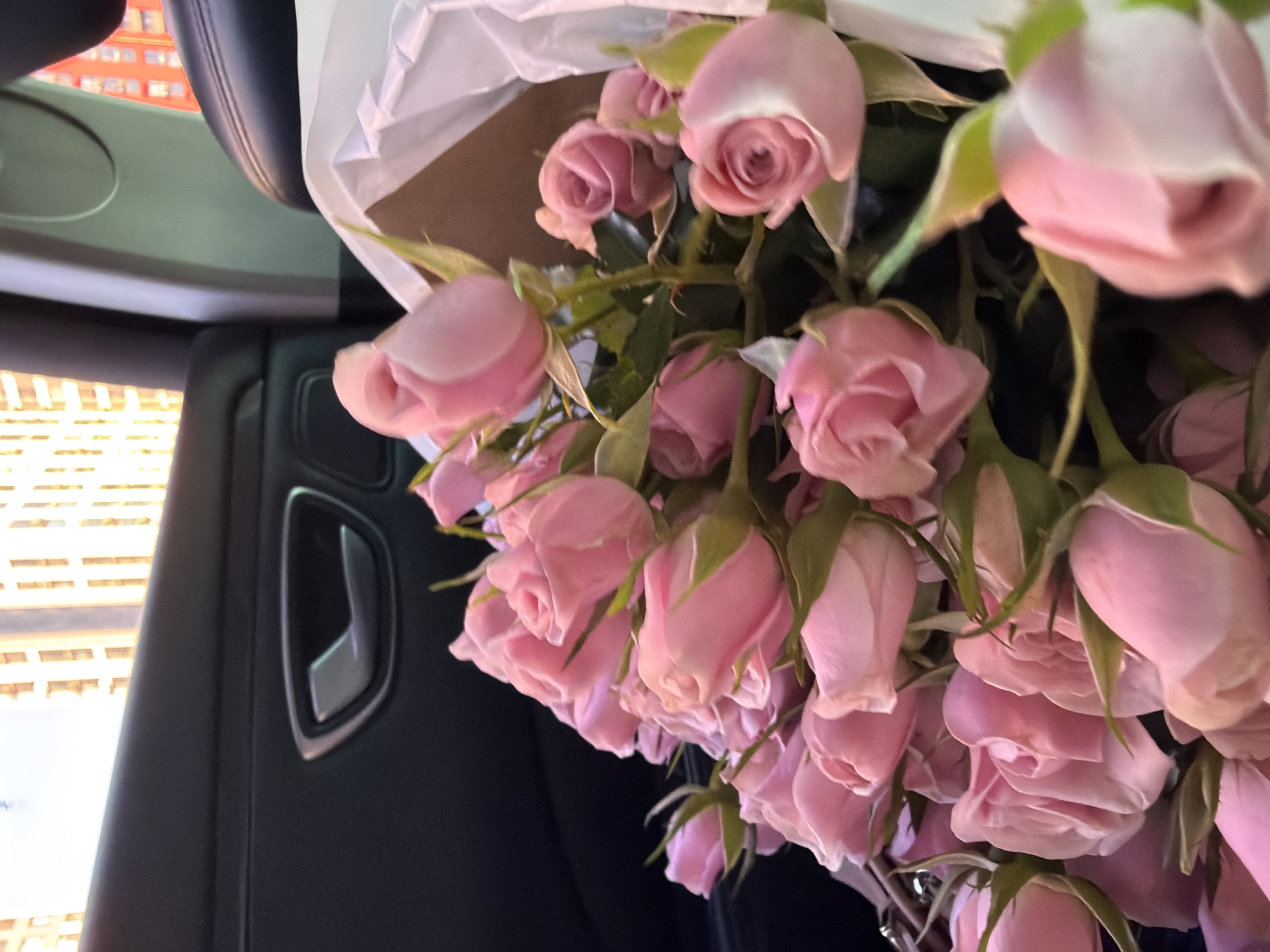 A bouquet of pink roses is placed on the passenger seat of a car. The car's interior is visible, with a black leather door panel and a silver door handle. Outside the car window, a modern building with a grid-like facade can be seen, suggesting an urban setting. The context of Kyiv, Ukraine, is provided for this scene.