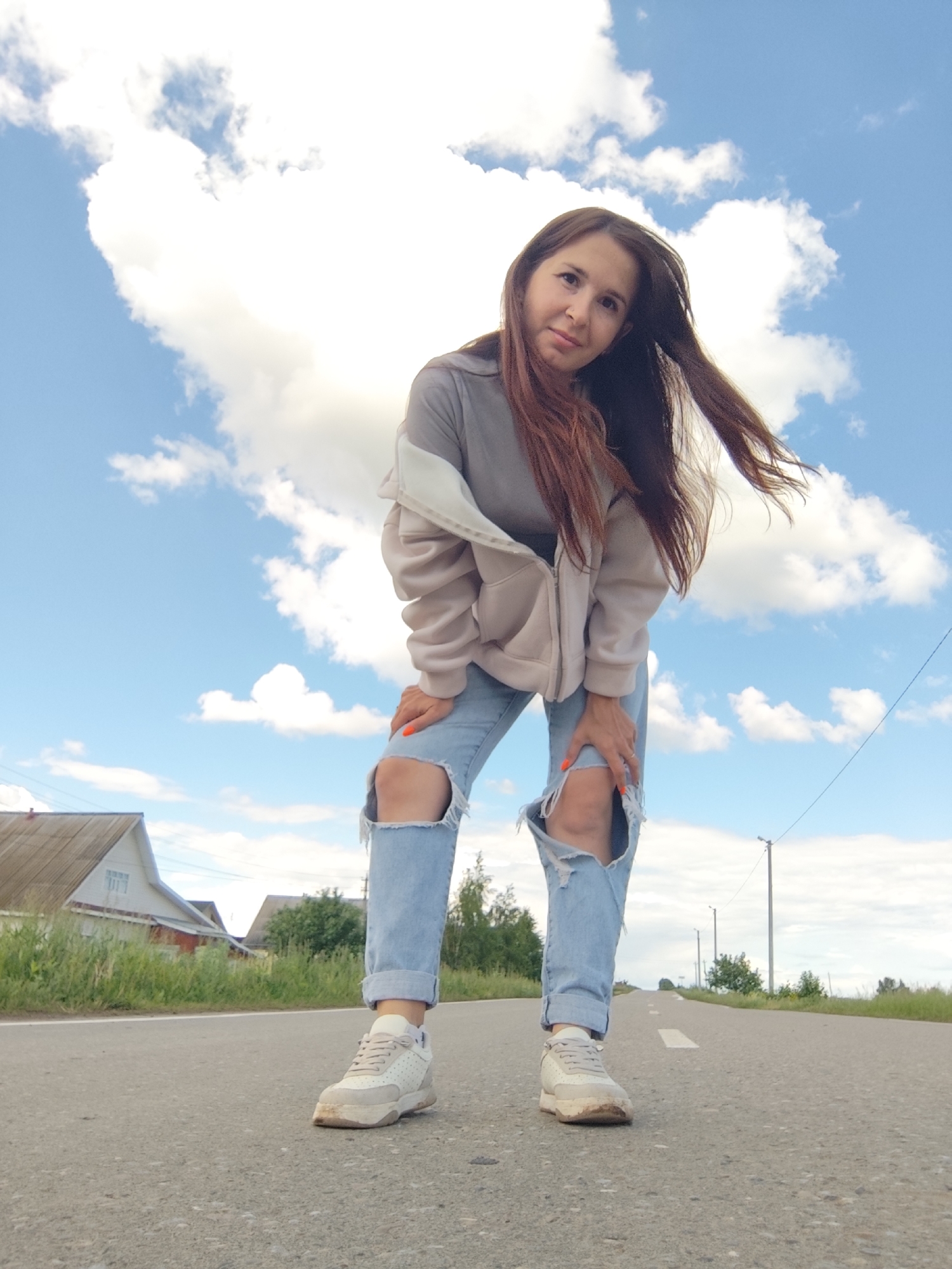A young woman stands on a paved road in Фоки, Russia, posing for a photograph. She is wearing ripped blue jeans, a grey t-shirt, and a light brown hoodie. Her brown hair is windblown. The road has a white painted line down the center. A house is visible on the left, and utility poles line the road in the distance. The sky is blue with fluffy white clouds.