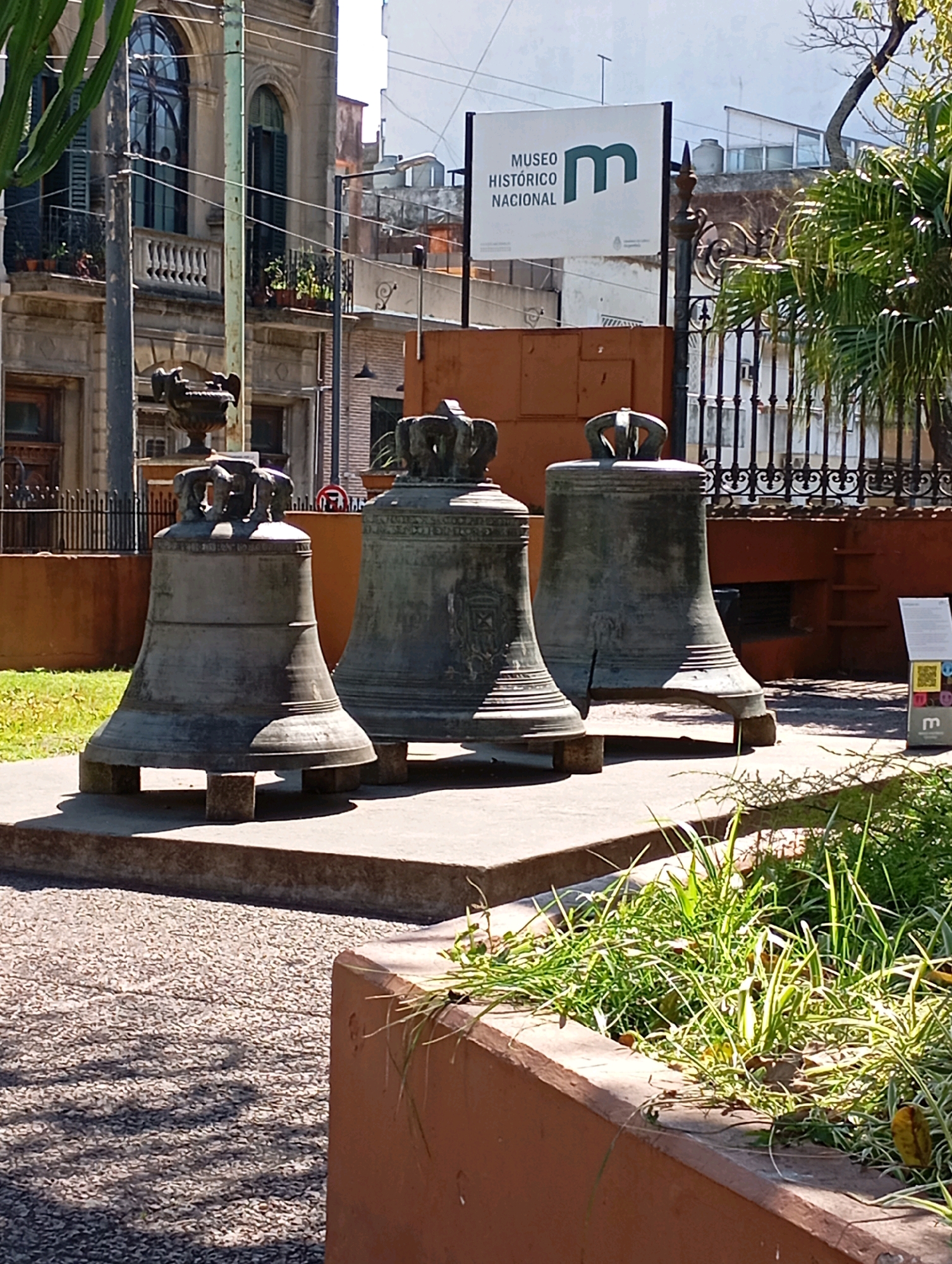 The image depicts an outdoor scene in Buenos Aires, Argentina, specifically at the Museo Histórico Nacional. In the foreground, three large, weathered bronze bells are prominently displayed on concrete pedestals. The bells are ornate, with intricate designs and crowns at their tops. To the left of the bells, a tall, thin pole with electrical wires runs vertically. Behind the bells, a low, reddish-brown wall forms a boundary, and beyond that, a black wrought-iron fence separates the museum grounds from the street. A sign with the museum's name and logo is visible behind the fence. In the background, a multi-story building with arched windows and balconies can be seen, suggesting the historical architecture of the area. Lush green grass and foliage are present in the foreground and to the right, with sunlight casting shadows on the ground, indicating a daytime setting. There are no people visible in the scene.