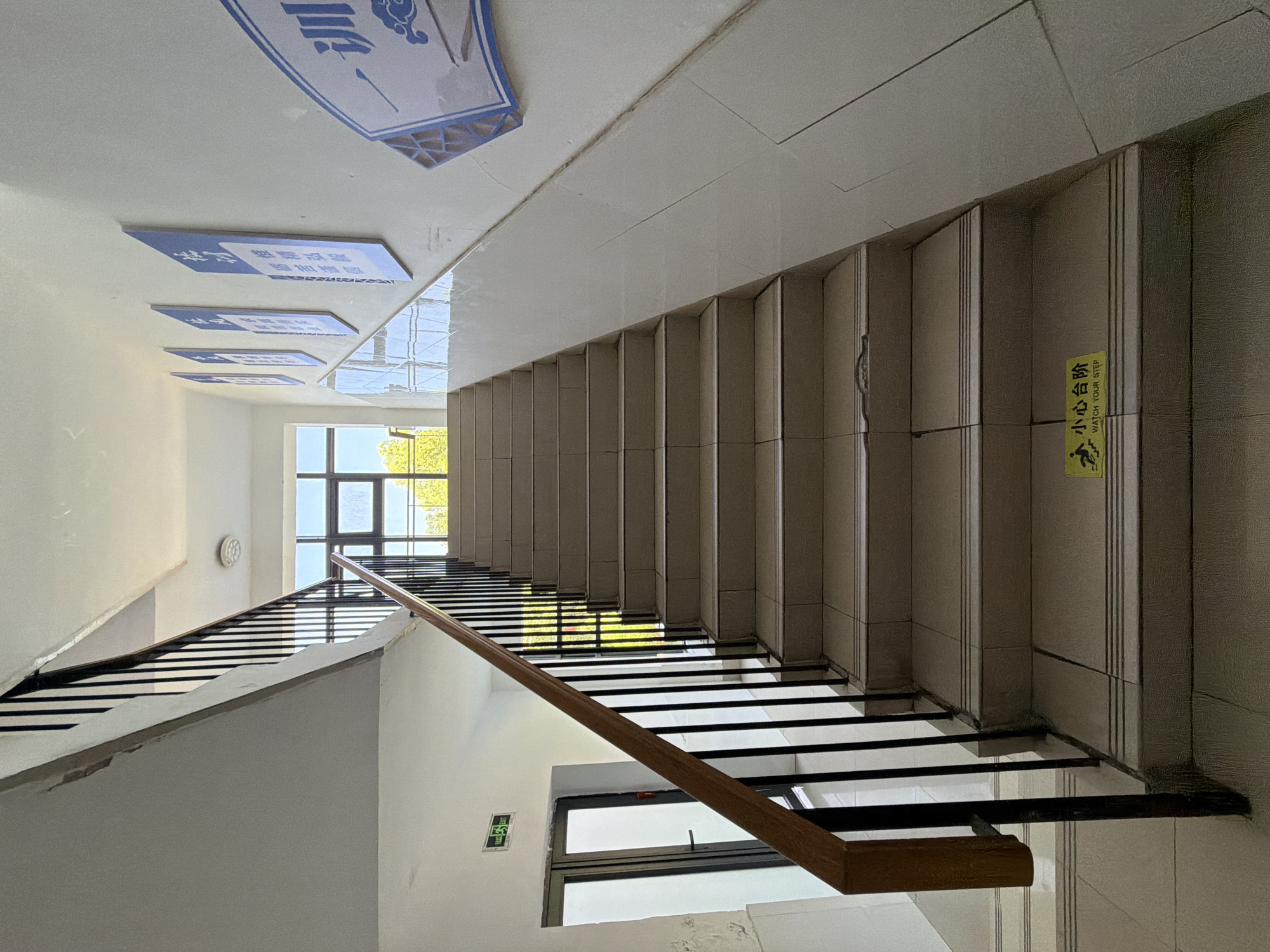 The image shows an indoor staircase in what appears to be a public building, possibly a school or administrative facility, located in Jiangjin, China. The staircase has a tiled surface and a black metal railing with a wooden handrail. On the wall to the right of the staircase, several blue and white signs with Chinese characters are mounted. A yellow sign with the text "小心台阶" and "watch your step" is affixed to one of the lower steps. To the left of the staircase, there is a door with glass panels and a green "EXIT" sign above it. Natural light streams in from a window at the top of the staircase, illuminating the area. The overall impression is one of an ordinary, functional interior space.