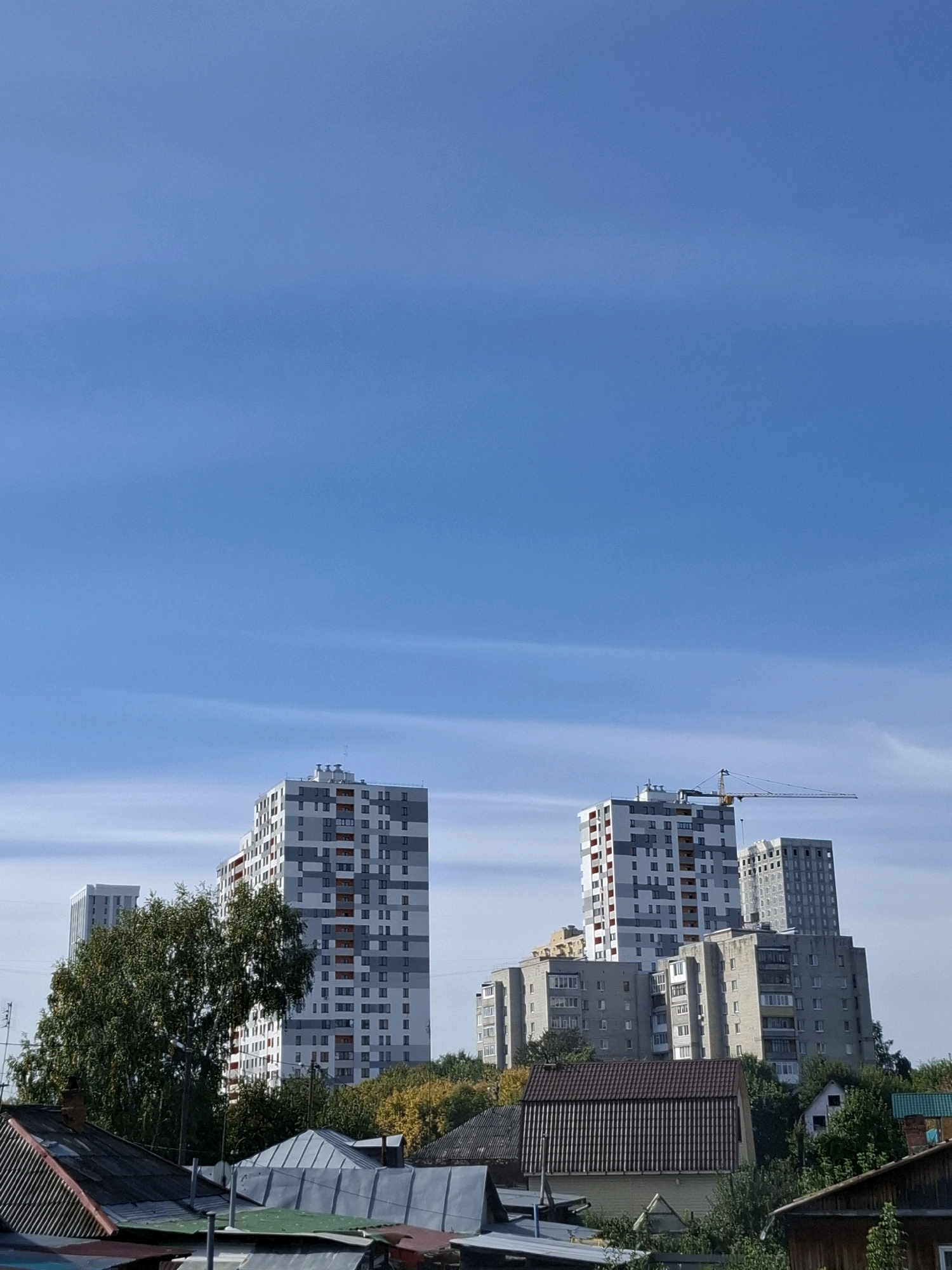 The image depicts a cityscape with several multi-story buildings under a clear blue sky. In the foreground, there are rooftops of smaller structures, possibly residential houses with corrugated metal roofs. Behind these, a lush green tree stands prominently, partially obscuring a tall, modern apartment building with a white and grey facade and red accents. To the right of this building, another, slightly shorter apartment building is visible, with a construction crane extending over it, suggesting ongoing development. Further in the background, more apartment complexes can be seen. The scene is typical of an urban environment undergoing construction and development, with a mix of older and newer structures. The geographical location is the city of Tyumen, Russia.