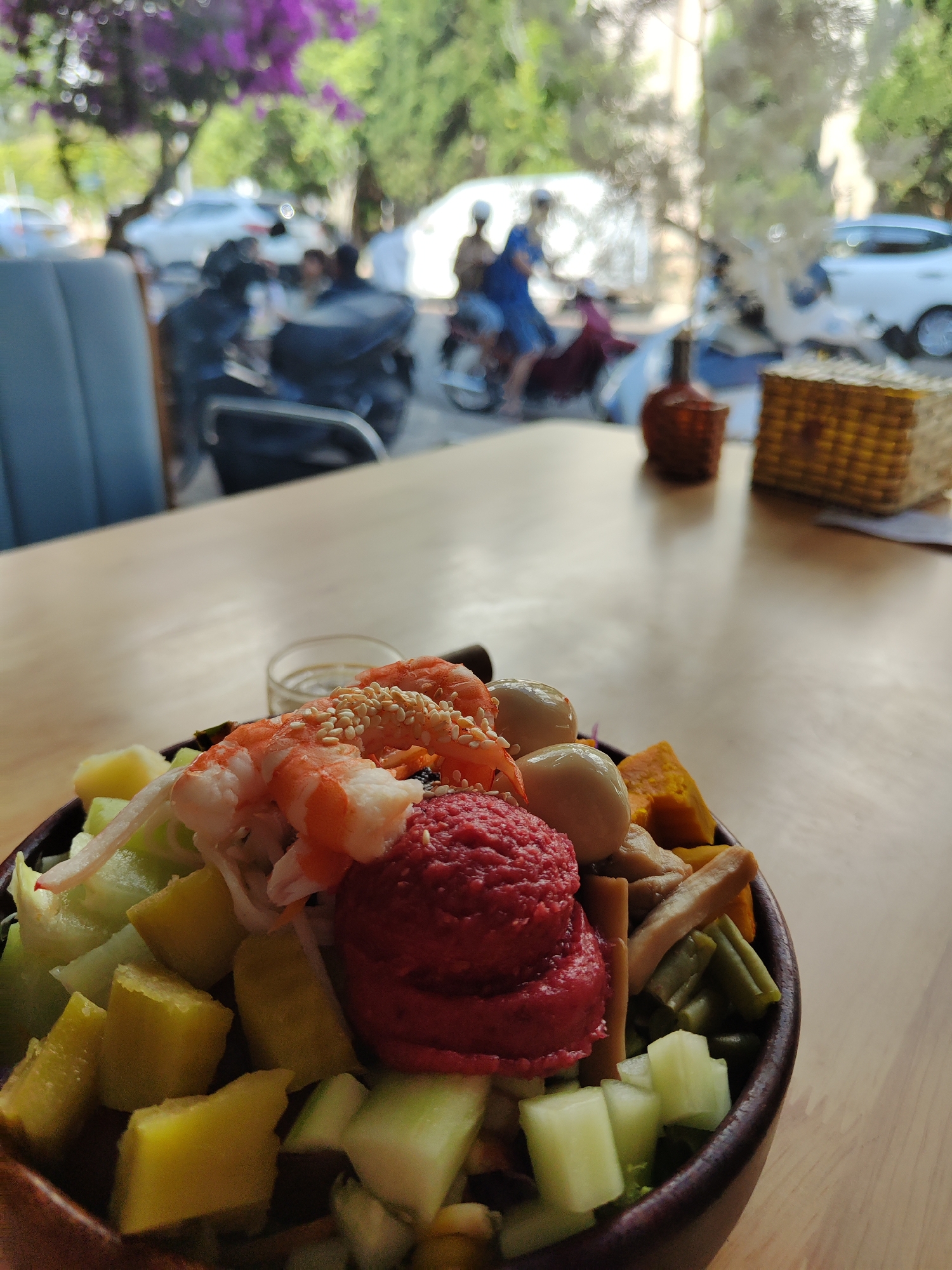 A vibrant salad bowl takes center stage on a wooden table inside a restaurant, with a blurred cityscape visible through the window behind it. The bowl is filled with a colorful array of ingredients, including:
* Cooked shrimp sprinkled with sesame seeds
* Red beet puree
* Diced mango and cucumber
* Quail eggs
* Green beans
* Cubed pumpkin
* Bamboo shoots
In the blurred background, the window reveals a street scene in the city of Da Lat, Vietnam. One can identify parked motorbikes and a couple of people on a scooter driving past a building with purple flowers in front of it. The background features a light blue cushioned chair, a brown decorative bottle, and a small yellow woven basket on the table. The overall scene suggests a casual dining environment.
