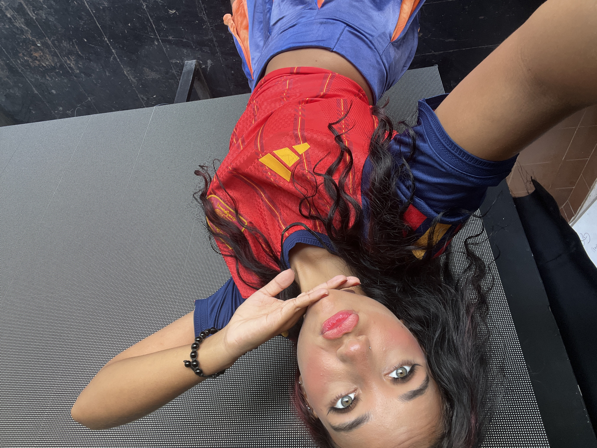 A young woman is lying on her back on the floor, looking up at the camera and making a kissing face. She has dark, curly hair with streaks of pink, light green eyes, and is wearing a red and blue Adidas soccer jersey and a blue and orange skirt. She has a black beaded bracelet on her left wrist and a nose ring. Her right arm is bent at the elbow, with her hand resting on her chin. The background is a large, grey, textured surface with a pattern of small white dots, possibly a wall or a large screen. To the left of the woman, a black object, possibly clothing, is visible, and to the far left, a light-colored surface with some writing can be seen. The floor beneath her is dark and appears to be made of wood or a similar material with a distressed finish. The image appears to be a selfie taken indoors. The presence of the grey dotted surface and the wooden floor suggest a modern interior space, potentially an event venue, studio, or a home with a specific design aesthetic. Without further context, it's impossible to definitively place this scene within a specific location in Porlamar, Venezuela.