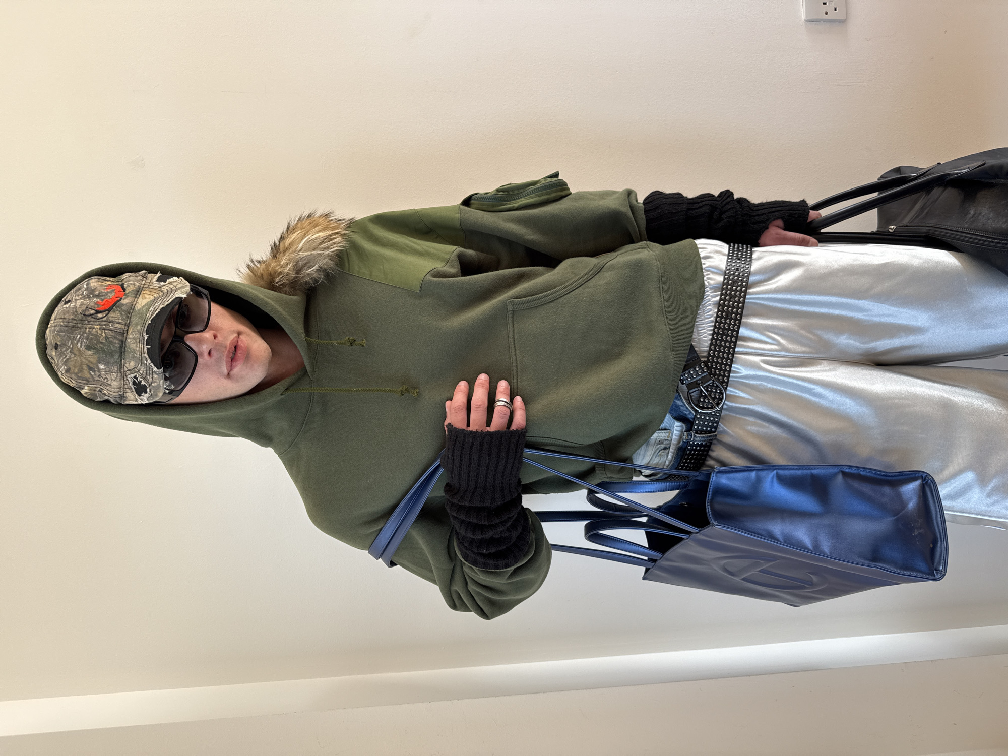 A young person stands indoors, posing for a photograph. They are oriented slightly to their left, dressed in a distinctive casual outfit, against a plain light beige wall. A white electrical outlet is visible on the wall in the upper right background.
The individual wears a camouflage baseball cap with an orange logo, partly covered by the raised hood of a dark olive green hooded sweatshirt. Dark sunglasses obscure their eyes. The hoodie features a brown fur trim around the hood's opening, suggesting warmth. On their arms, they wear black knitted fingerless arm warmers.
Below the sweatshirt, the person has on shiny silver or metallic-looking trousers. These are cinched at the waist with a black belt adorned with numerous silver studs and a prominent dark buckle. They are holding two bags: a dark blue rectangular tote bag, likely made of leather, with the embossed 'T' logo of the brand Telfar clearly visible on its side, held in their left hand. A larger, black duffel bag or travel bag with black handles is held in their right hand. A silver ring is visible on the ring finger of their left hand.
The person is looking directly towards the camera with a neutral expression. The scene is a simple indoor environment, possibly a room or hallway. No specific London cues are visible in the image itself.