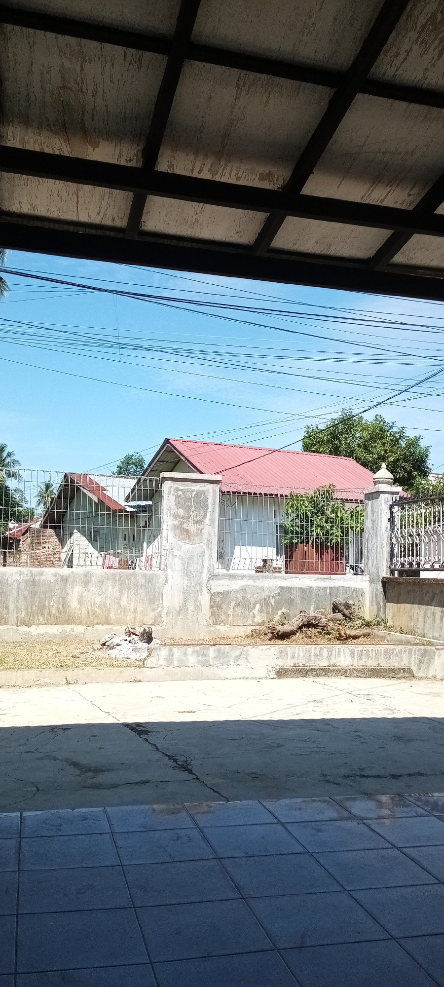 The image, taken from under a covered area, depicts a street scene in Lhokseumawe, Indonesia. Across the street is a property enclosed by a concrete and wire fence. Two houses are visible behind the fence; one is lighter in color with a red roof, and the other appears to be made of brick. Some logs and debris lie on the ground between the fence and the road. Power lines are visible against a clear, blue sky. The foreground shows tiled flooring beneath the point of view, with a mix of light and shadow.