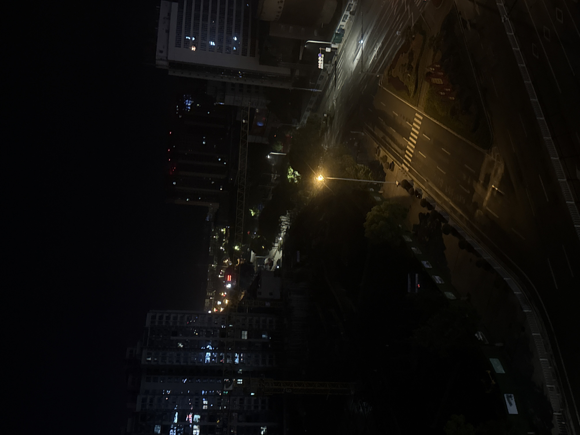 The image shows an elevated nighttime view of a city street in Zhangjiajie, China. A tall building with illuminated windows is visible to the left and slightly above, partially shrouded in darkness. A street lamp provides light to a road below, revealing a zebra crossing. Dark greenery is situated near the road, and vehicles can be seen parked on the side. In the lower left, another building with illuminated windows and a partially visible crane. The rest of the scene is enveloped in darkness.