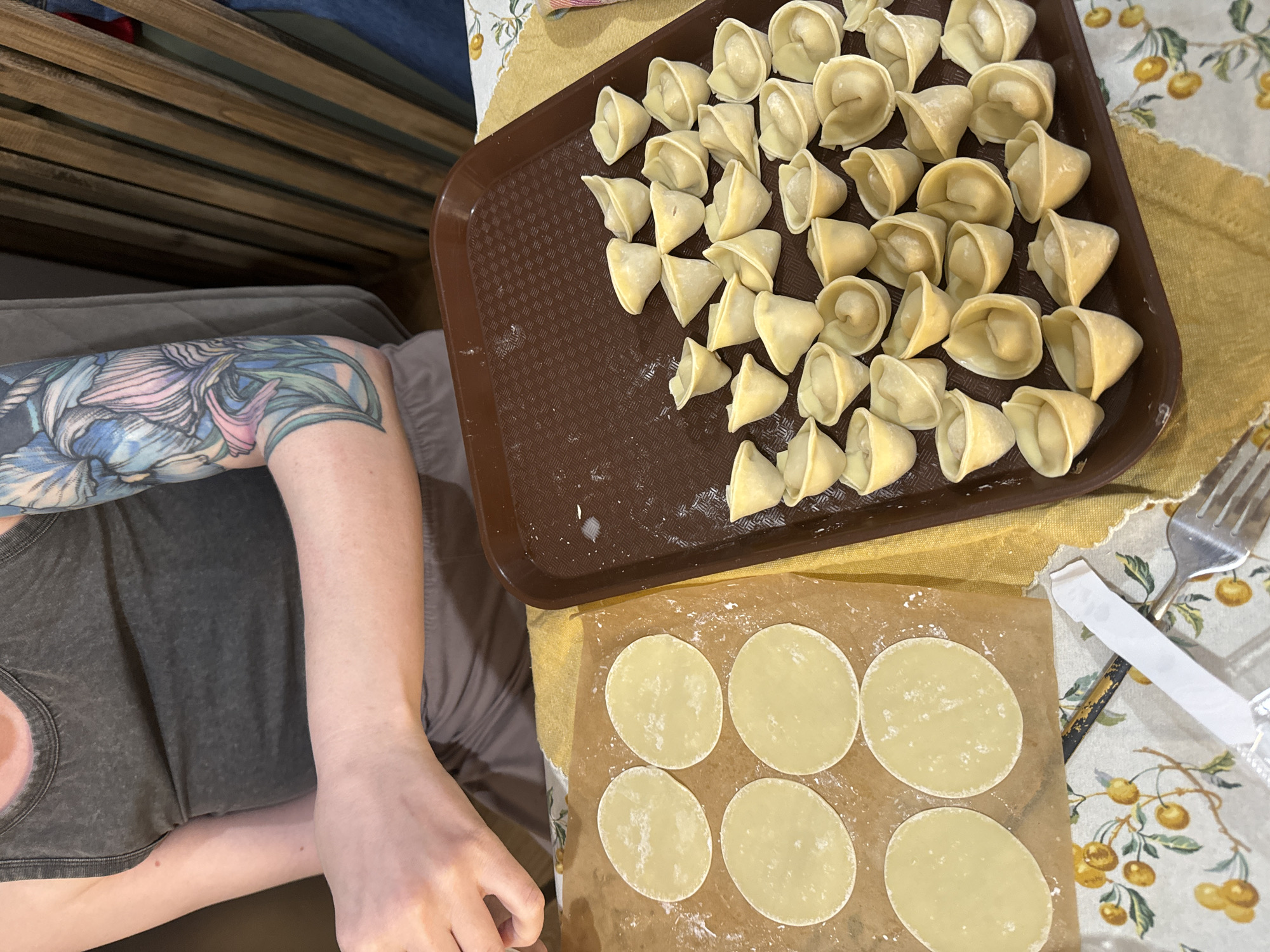 A medium-angle, eye-level shot shows a person with a floral tattoo on their left forearm sitting at a table making pelmeni (dumplings). The person is wearing a gray tank top. A brown tray full of pelmeni sits on a yellow and white patterned tablecloth. Beside the tray, dough circles are laid out on parchment paper. A fork and what appears to be a dumpling press rest on the tablecloth as well. The scene is likely taking place indoors in Krasnoyarsk, Russia.