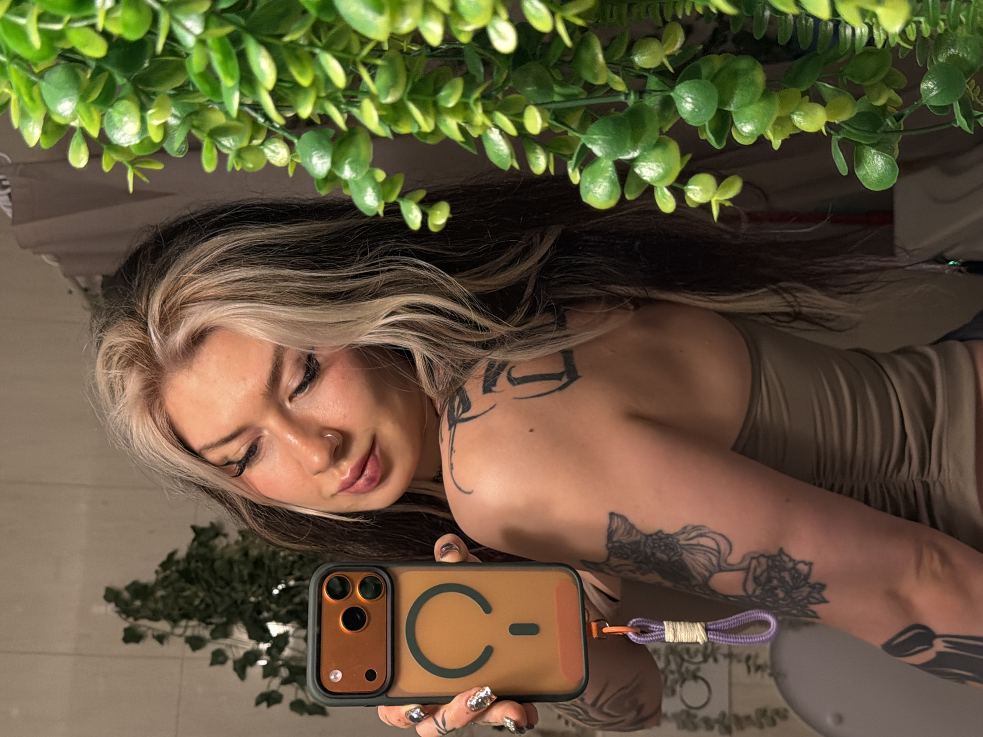 A woman with blonde-streaked brown hair is reflected in a mirror, holding up a cell phone with a brown and black case featuring a stylized "C-" design. She wears a beige top and has visible tattoos on her arm and shoulder. The background includes green foliage both above and below her reflection. The location is not identifiable in the image.