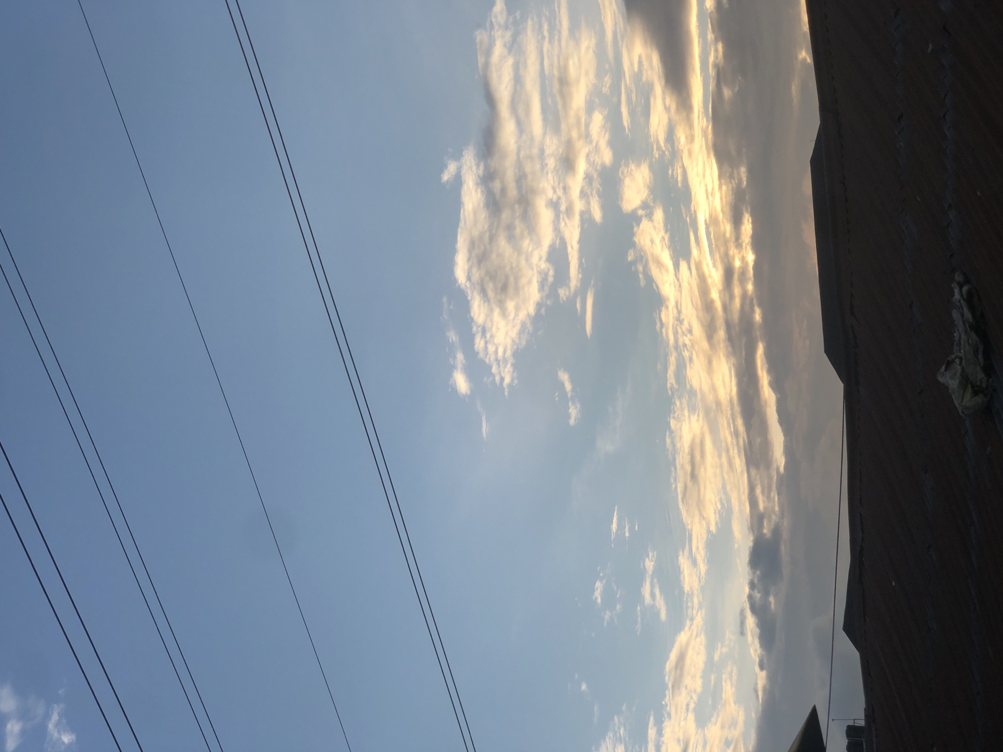 The image captures a view of the sky and rooftops in Ado, Nigeria. Several dark utility lines are stretched horizontally across the upper portion of the frame against a pale blue sky. Below the sky, scattered clouds are visible, tinged with the warm hues of sunset or sunrise, suggesting an early morning or late evening time. The lower third of the image is dominated by the dark, corrugated tin rooftops of several buildings. One building in the upper left corner has a visible water tank on its roof. The overall scene is urban and suggests a residential area.