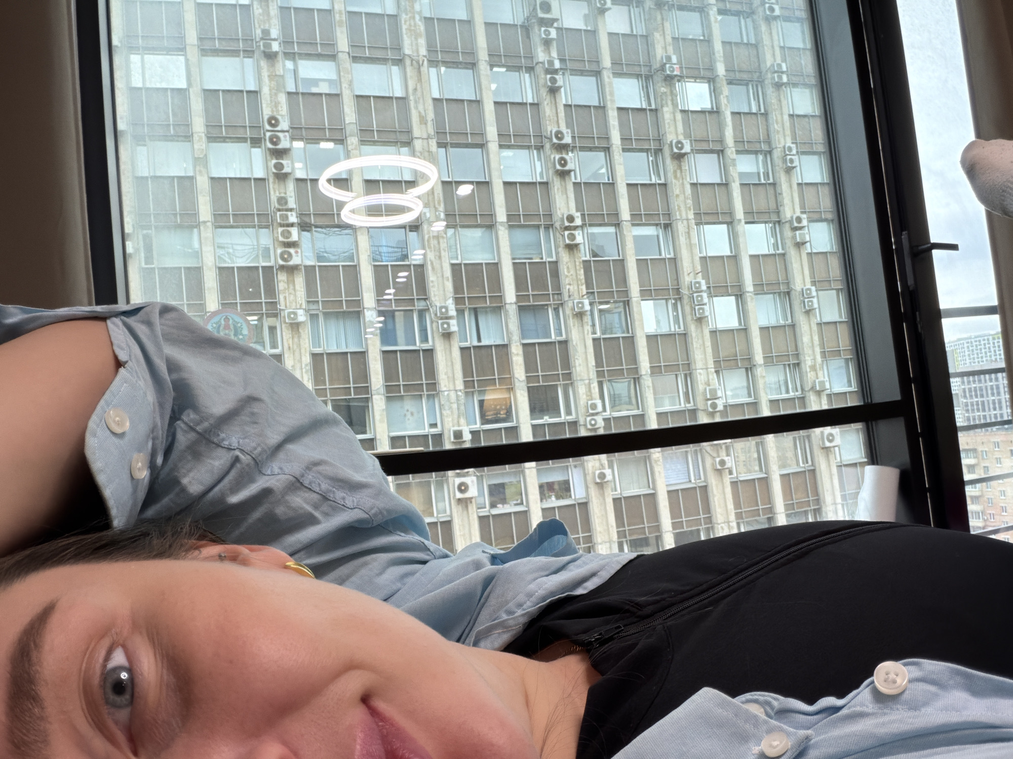 A woman is lying down indoors, with a large building visible through a window behind her. The building is tall with a repetitive pattern of windows and air conditioning units, a common architectural style in Moscow. There are two decorative halo-shaped light fixtures hanging indoors near the window. The woman is wearing a light blue button-down shirt and black clothing underneath. The environment suggests an urban setting. The image does not provide definitive proof that it is taken in Moscow, but the architectural style of the building is characteristic of that city.