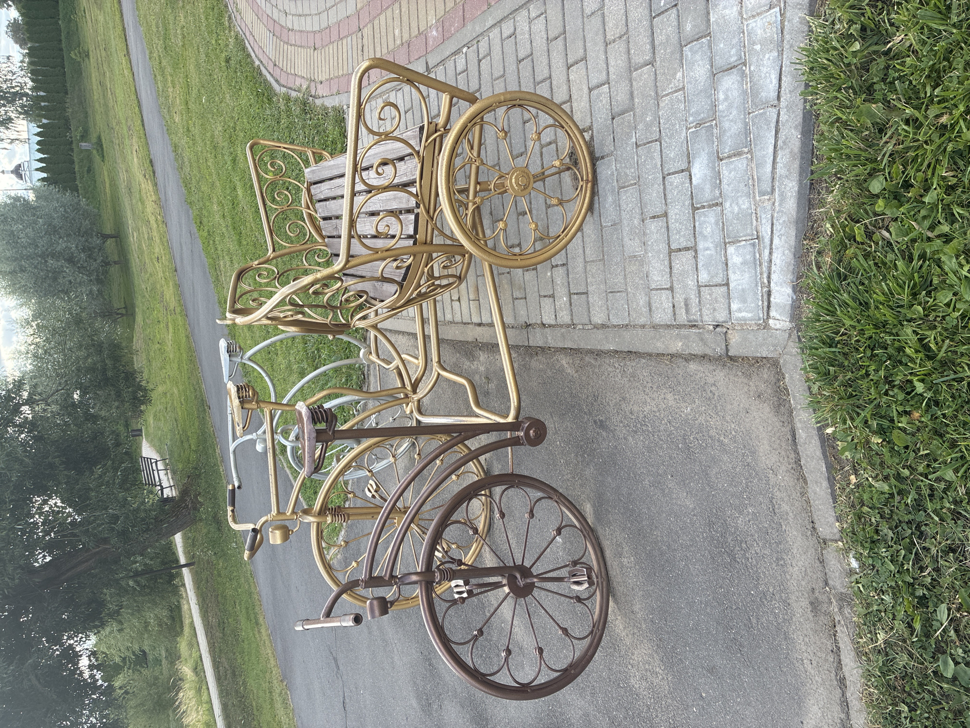 The image displays a decorative, two-tiered metal bicycle-shaped bench positioned on a paved walkway adjacent to a grassy area. The bench is ornate, with intricate scrollwork on the backrest and sides, and appears to be made of gold and bronze-colored metal. The seating area is constructed from wooden slats. Behind the bench, two other metal bicycle-themed decorations are visible, one predominantly gold and another in a lighter, possibly silver or white hue. The background features a paved path winding through a green, well-maintained park with trees and grass. In the distance, a white building with a dome, possibly a church, can be seen. The scene suggests a public park or garden setting in the city of Mahilyow, Belarus, given the architectural style and general ambiance. There are no people or animals visible in the frame.
