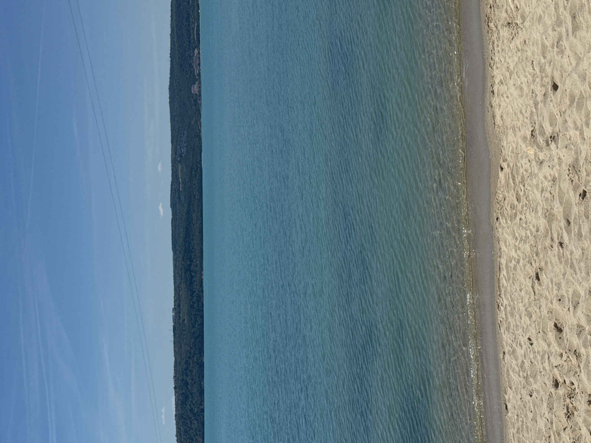 The image shows a beach scene in Varna, Bulgaria. A sandy beach stretches to the right side of the frame. Clear, turquoise water extends towards the left, merging with the shoreline. On the opposite shore, a dense, green forest lines the coast. The sky above is clear and blue with faint contrails. Power lines can be seen stretching across the sky.