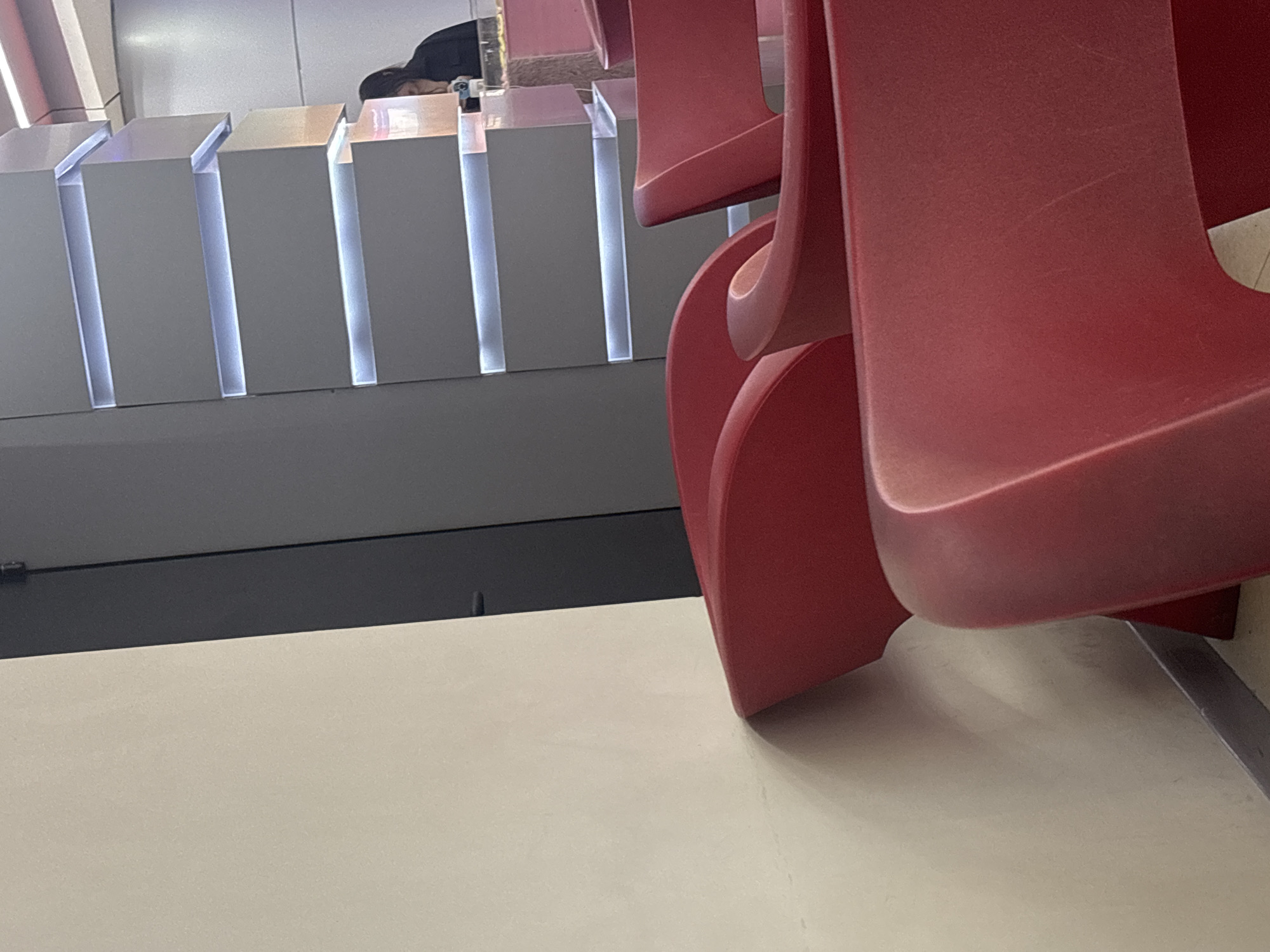 In an indoor setting within Yerevan, Armenia, the image presents a modern and somewhat austere public area. Dominating the right side of the frame are several red, contemporary chairs, likely made of plastic, their curved forms catching the light. Visible scuff marks on their surfaces suggest regular use. On the left, a series of light gray, rectangular modules with vertical, bright white LED strip lighting create a distinct, modern aesthetic, possibly forming part of a reception desk or a display counter. Behind these illuminated structures and a subtle transparent partition, a person with dark hair is seen resting, likely sleeping, with their head down. The floor in the foreground is a smooth, light-colored surface, contrasting with a dark gray base panel that runs beneath the illuminated modules. The artificial illumination throughout the scene creates a quiet, almost sterile ambiance, characteristic of a waiting room, airport lounge, or a modern office lobby. There is no visible text in the image. The precise time of day is not evident due to the reliance on indoor lighting.