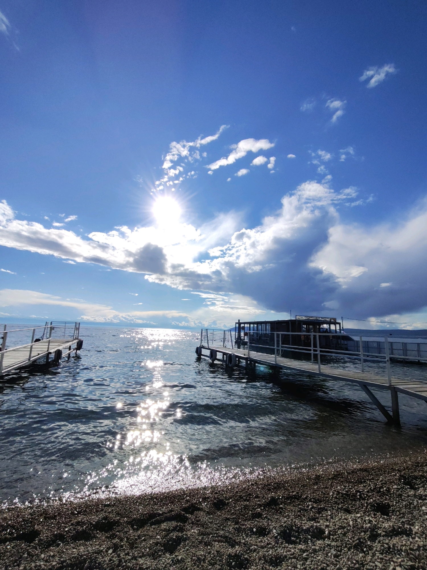 The image captures a serene lake scene under a bright, partly cloudy sky. Two wooden piers extend into the calm water, with the one on the right featuring a covered boat docked alongside it. The sun is high in the sky, casting a dazzling reflection on the water's surface. The foreground shows a pebble beach. The overall impression is one of a tranquil, sunny day by the water. This scene is characteristic of Listvyanka, a settlement on the southwestern shore of Lake Baikal, Russia, known for its picturesque lakeside landscapes and piers serving local boat traffic.