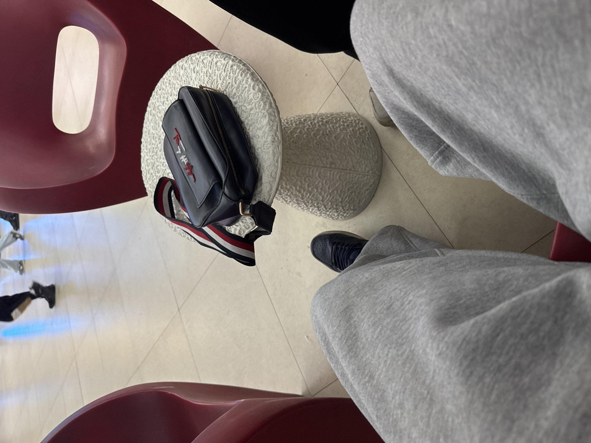 The image presents an overhead view of an indoor setting, likely a waiting area or lounge, in Yerevan, Armenia. The perspective is from a seated individual, whose legs, covered in grey sweatpants and dark shoes, are visible in the foreground and to the right.
Two modern, curved chairs in a reddish-maroon color flank the lower portion of the frame. Between these chairs and the seated person are two small, round, white textured stools or tables. A black handbag with a gold zipper and a distinctive red, white, and blue striped strap rests on one of these stools. The handbag features embroidered red text, clearly showing "TOMMY" followed by stylized letters, likely part of a brand logo.
The floor is made of light-colored, possibly beige or off-white, tiles with visible grout lines and some reflections, including a blue one in the mid-ground. In the background, another person is partially visible, walking away, wearing black shoes. The lighting suggests it is daytime. The overall atmosphere appears calm and casual.