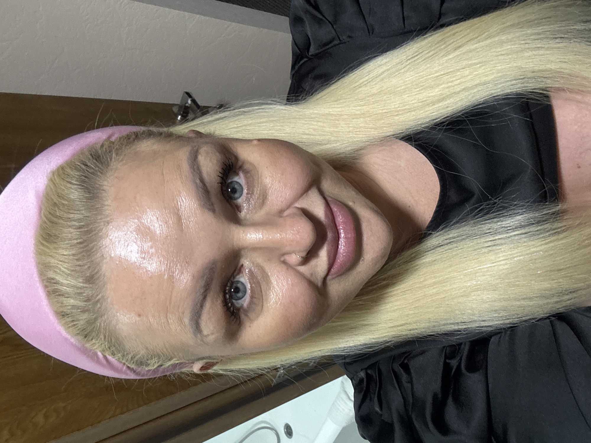 A headshot shows a woman with blonde hair and blue eyes, wearing a pink headband and a black top. Her skin appears shiny, possibly from sweat or makeup. She is looking directly at the camera with a slight smile. The background is blurred, showing what appears to be a wooden door on the left and a beige wall on the right. There are no discernible landmarks or indications of the city of Batumi, Georgia in the image.
